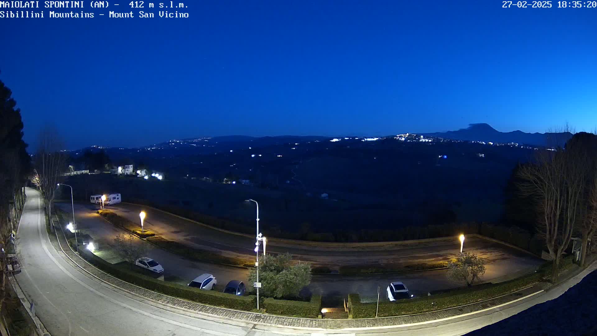 A curved road with parked cars is seen at dusk, overlooking a valley with distant lights under a clear, dark blue twilight sky.