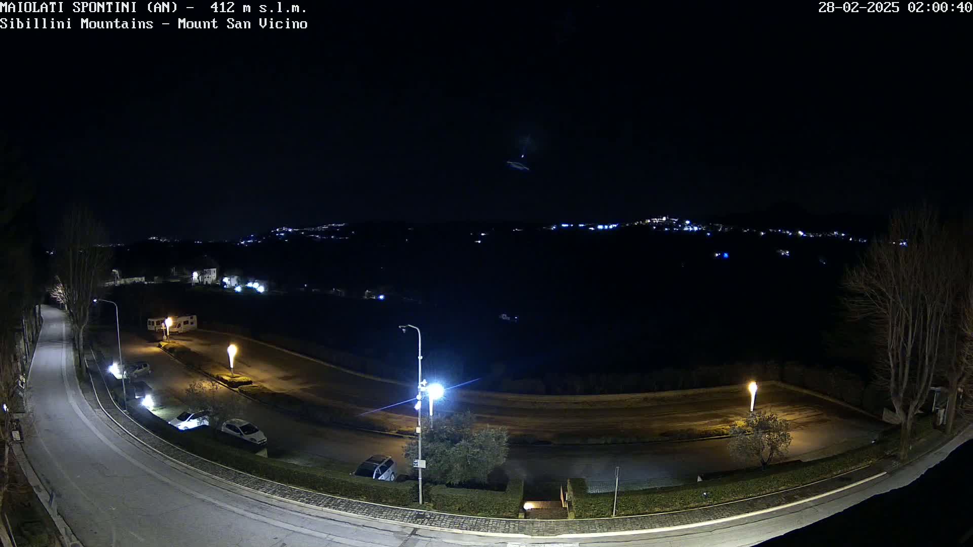 A nighttime, outdoor view of a curving road with parked cars and distant city lights under a clear sky.