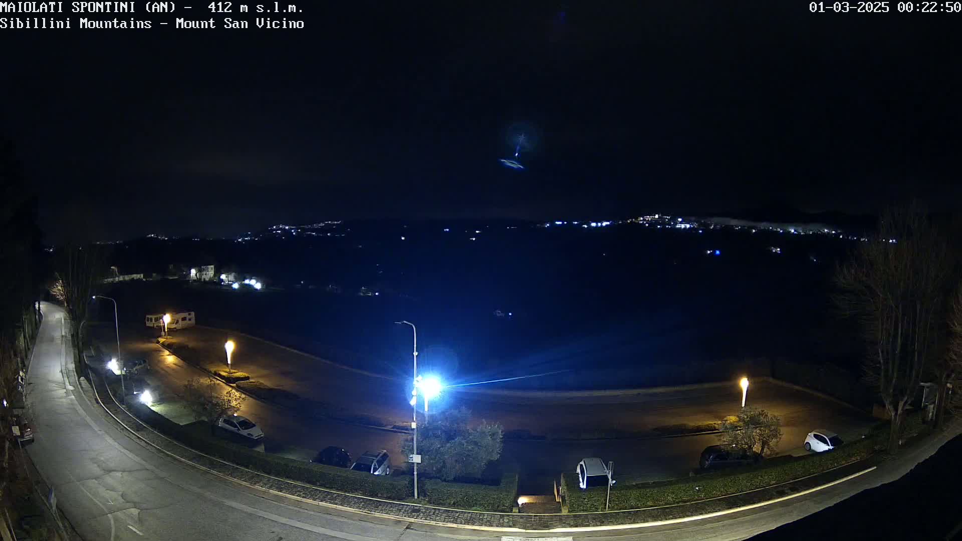 A nighttime, overhead view of a parking lot and curved road illuminated by streetlights, with a distant, faintly lit town and hills under a dark, clear sky.