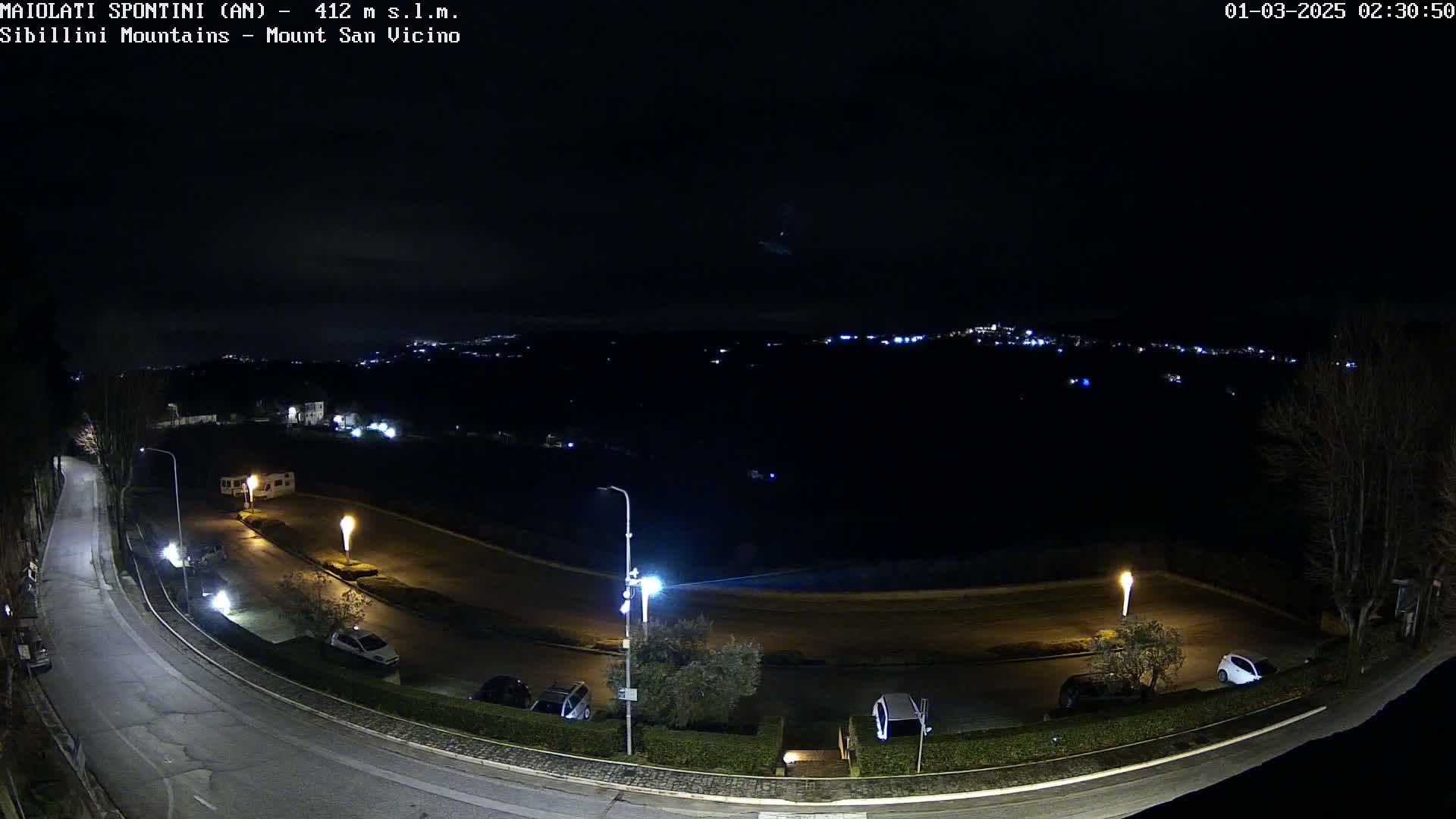 A nighttime, outdoor view of a curved road with parked cars and illuminated lampposts, overlooking a dark, distant town with faint lights under a clear sky.