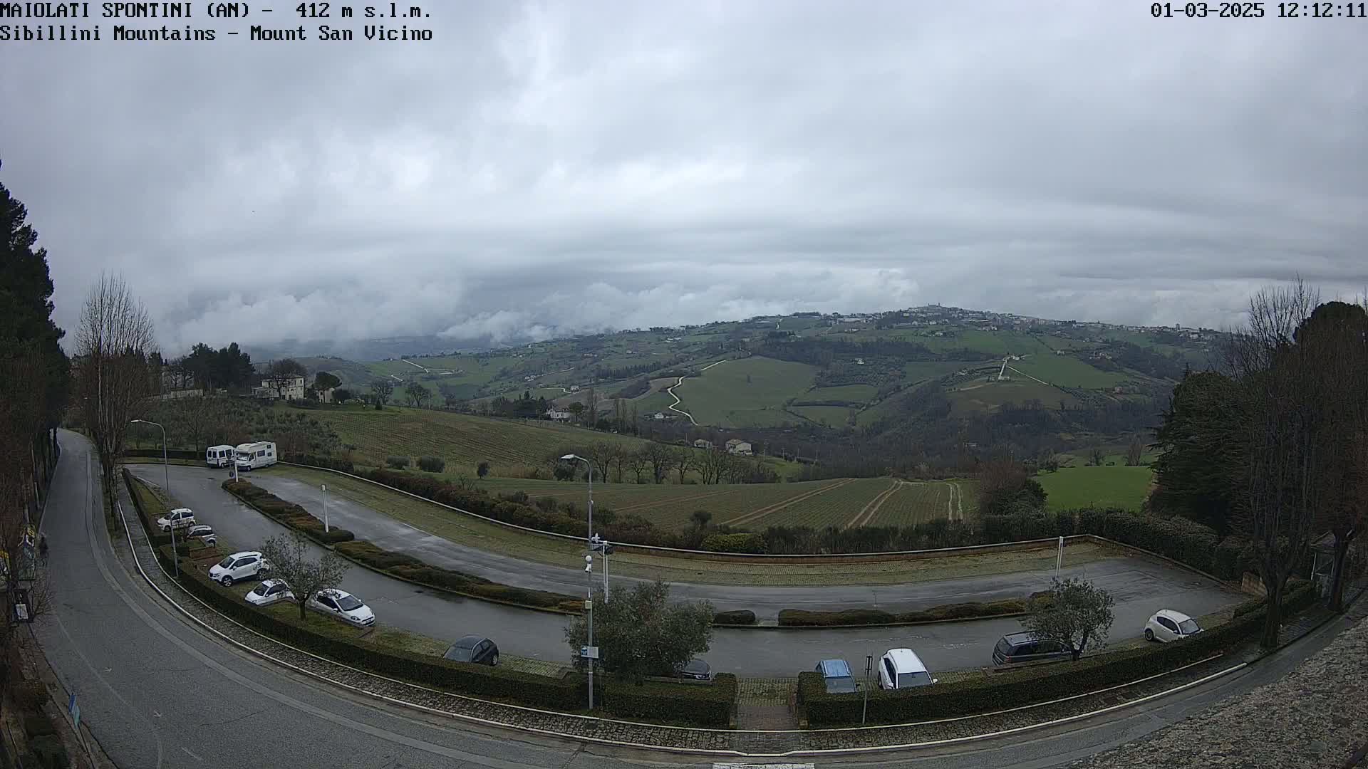 A paved road curves through a parking lot overlooking a valley of rolling green hills under an overcast sky.