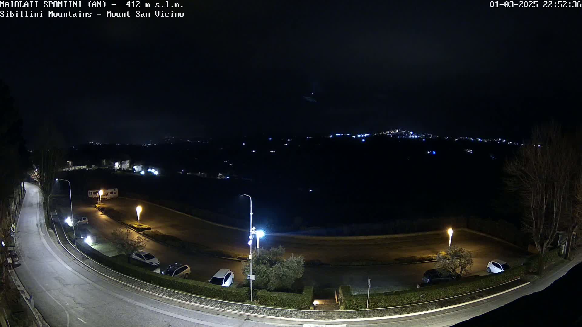 A nighttime, high-angle view of a curving road with parked cars, illuminated by streetlights, overlooks a dark, hilly landscape with distant city lights under a clear, dark sky.