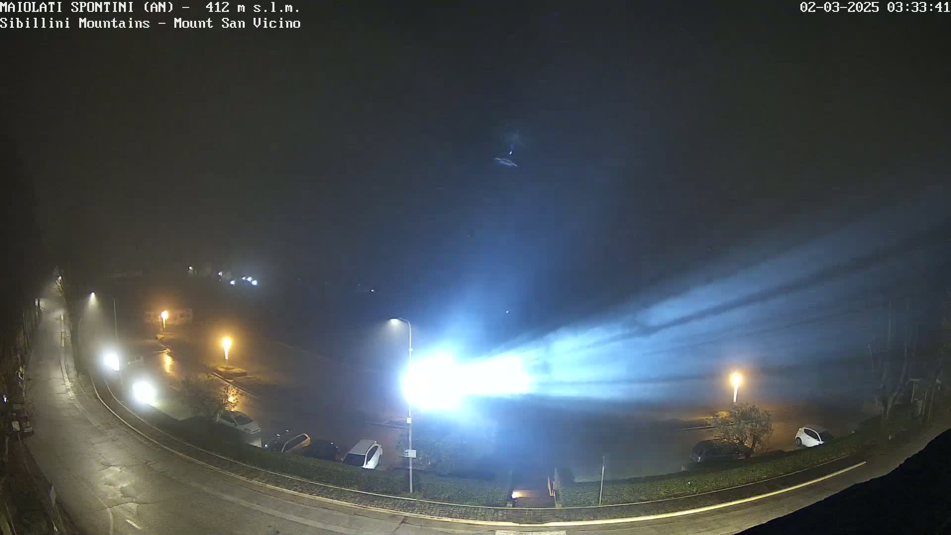 A nighttime, foggy scene shows a curved road with parked cars and streetlights, illuminated by beams of light cutting through the fog.