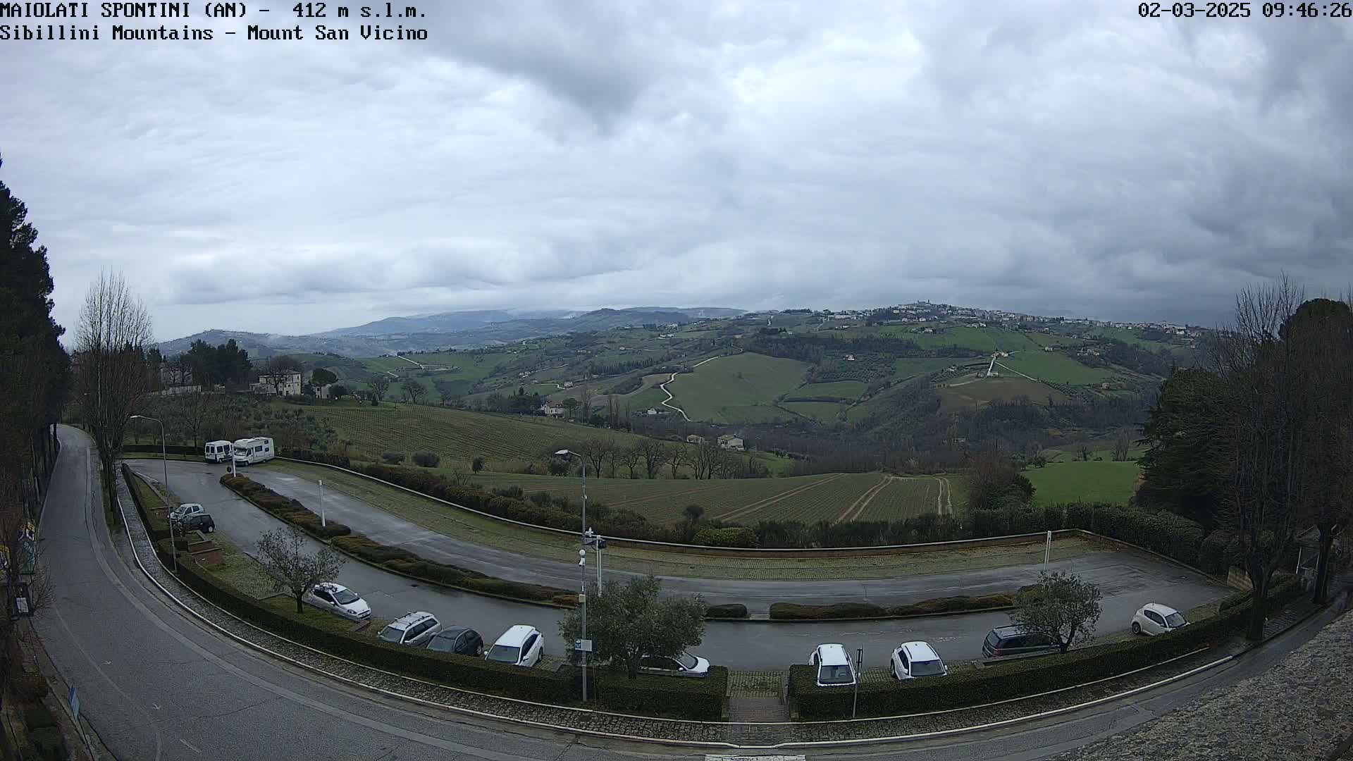 A parking lot with several cars and campervans overlooks a rolling green landscape under a cloudy sky.
