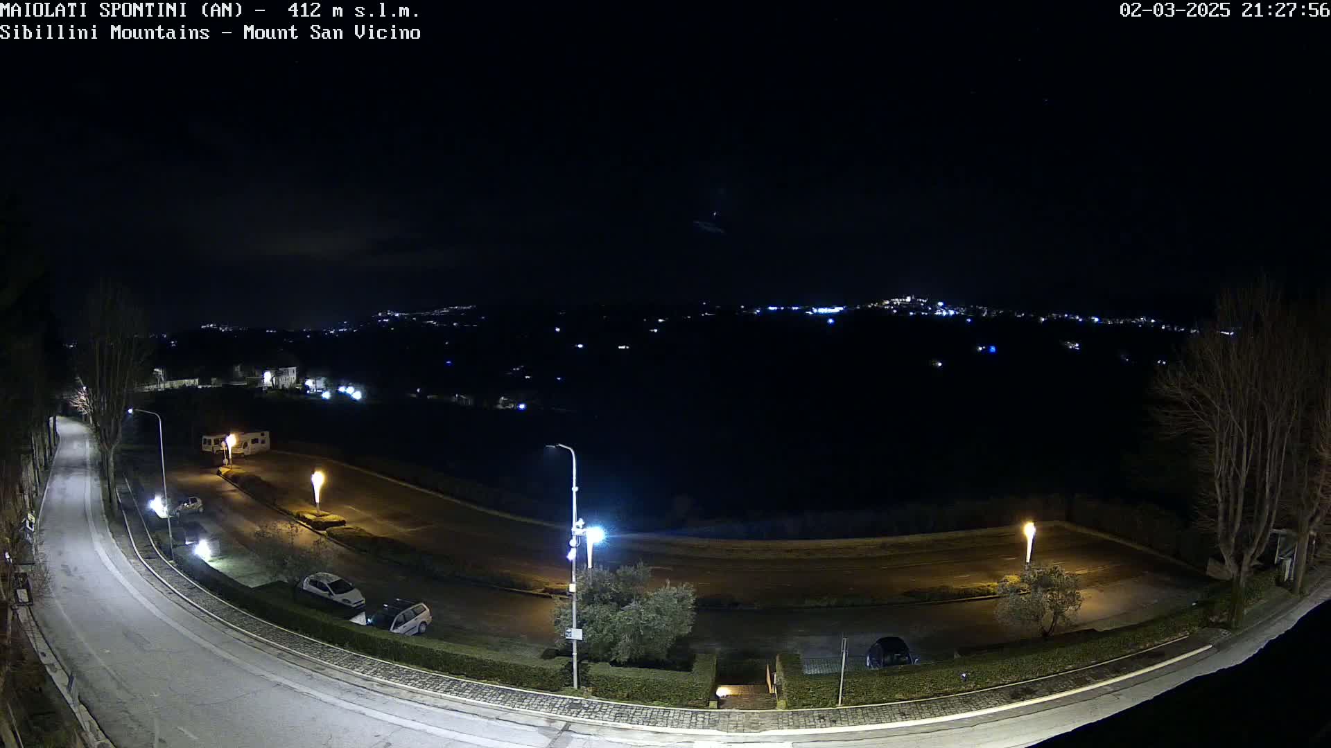 A nighttime, overhead view of a curving road with parked cars and streetlights, overlooking a distant town illuminated under a clear, starlit sky.