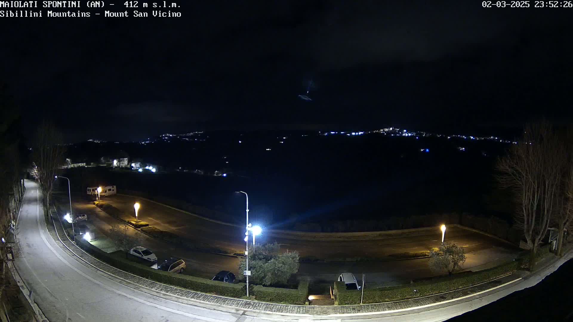 A nighttime, overhead view of a curving road with parked cars and streetlights, overlooking a distant town under a dark, cloudy sky.