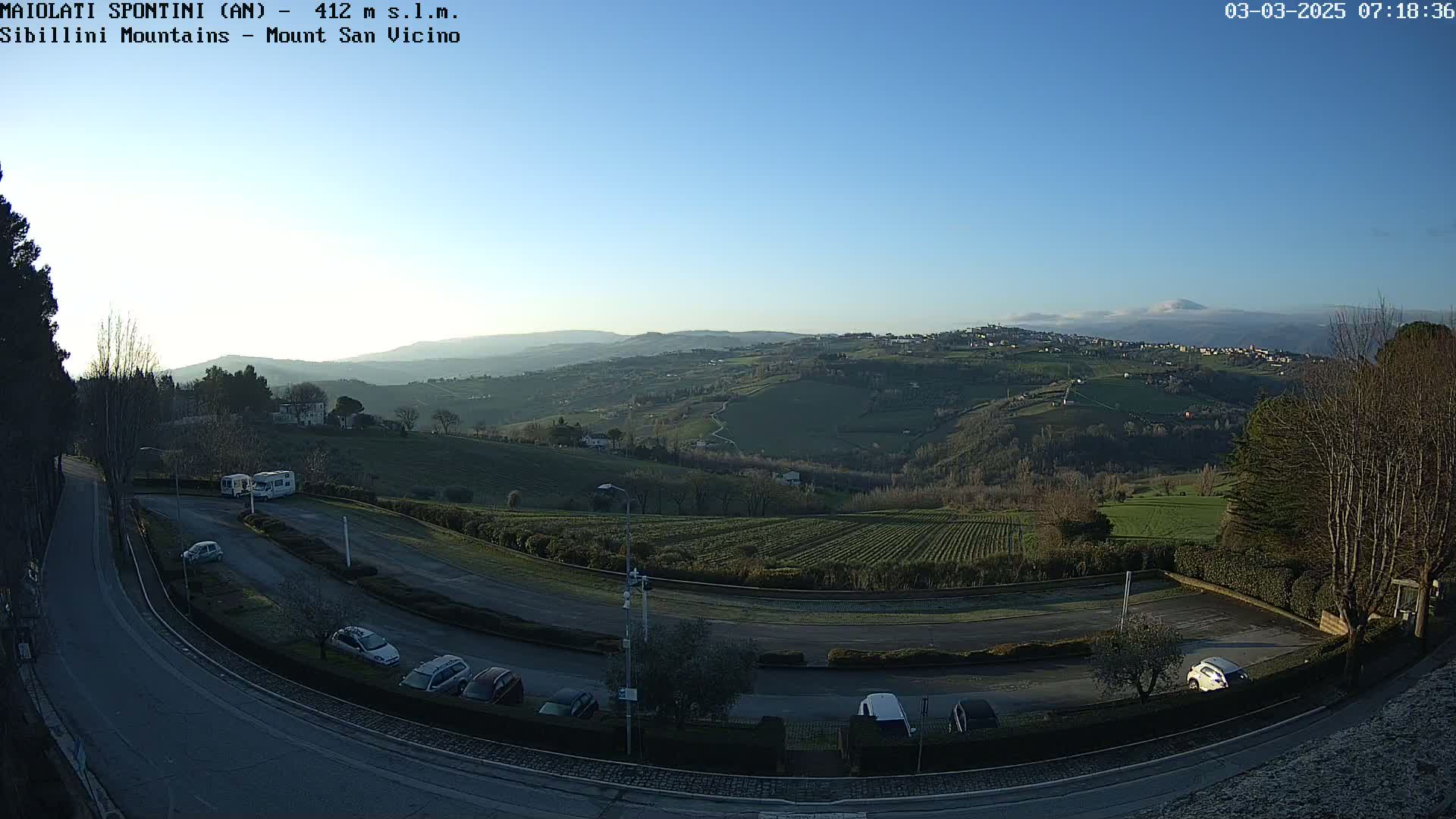 A clear, sunny morning view of a parking lot overlooking rolling hills and distant mountains, with several cars parked.