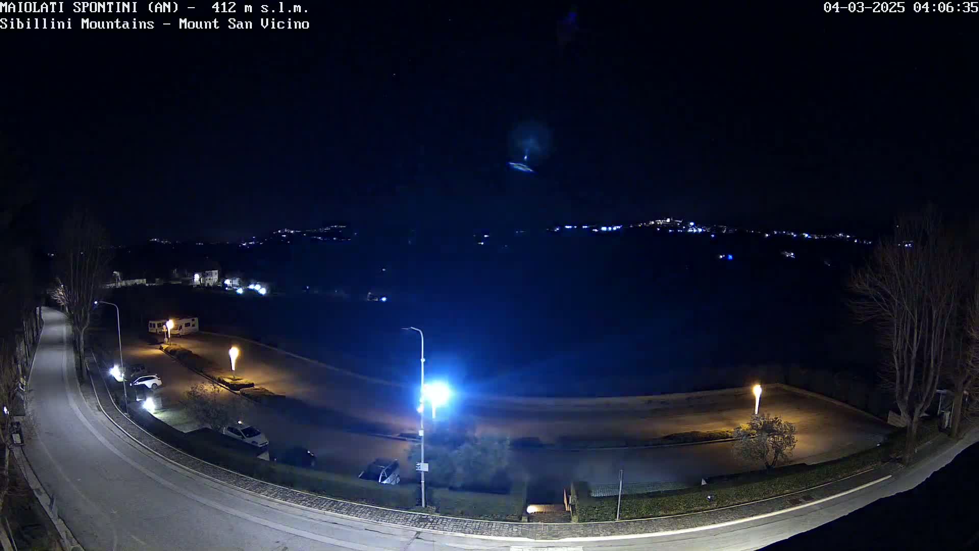 A nighttime, overhead view of a mostly empty parking lot and curving road, with a distant cityscape faintly visible under a clear, dark sky.