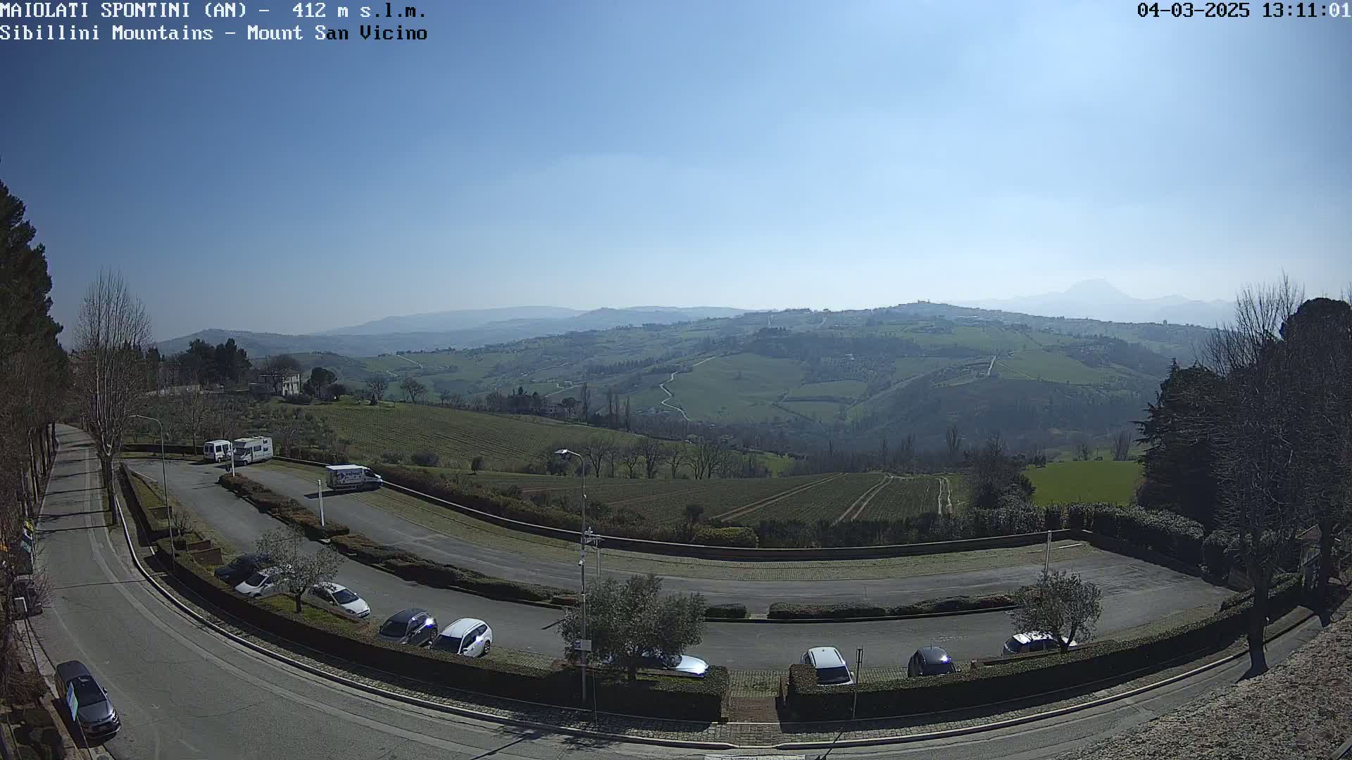 A sunny day overlooking a hilly landscape with vineyards and a parking lot containing several cars and campervans.