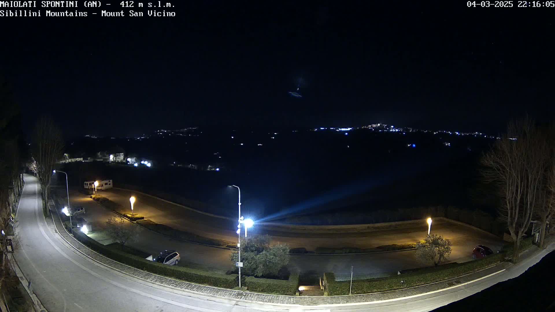 A nighttime, high-angle view shows a curving road illuminated by streetlights, with a few parked vehicles and a distant cityscape visible under a clear, dark sky.