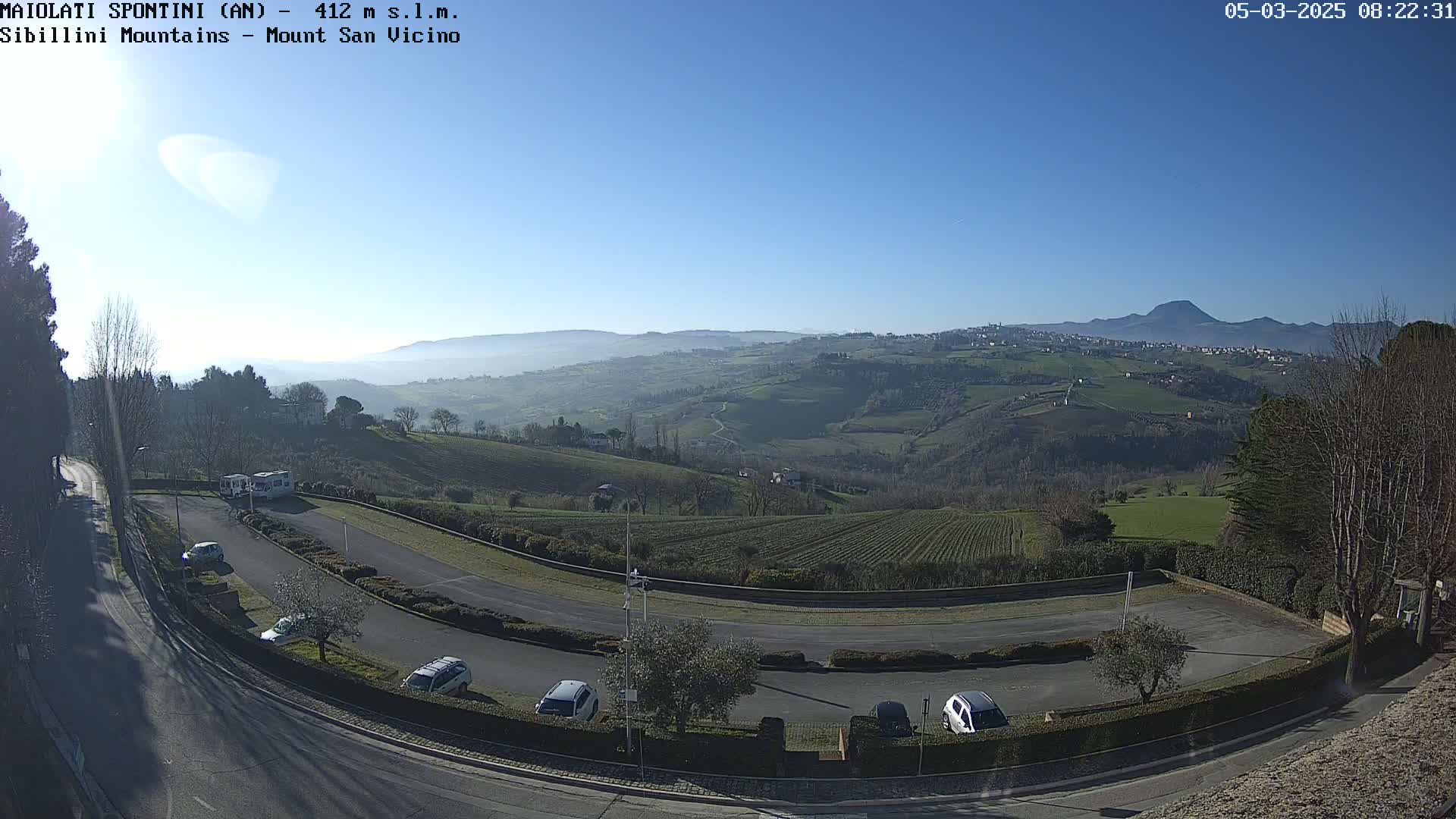 A clear, sunny day reveals a hilly landscape with a parking lot and several cars, overlooking a valley and distant mountains under a bright blue sky.
