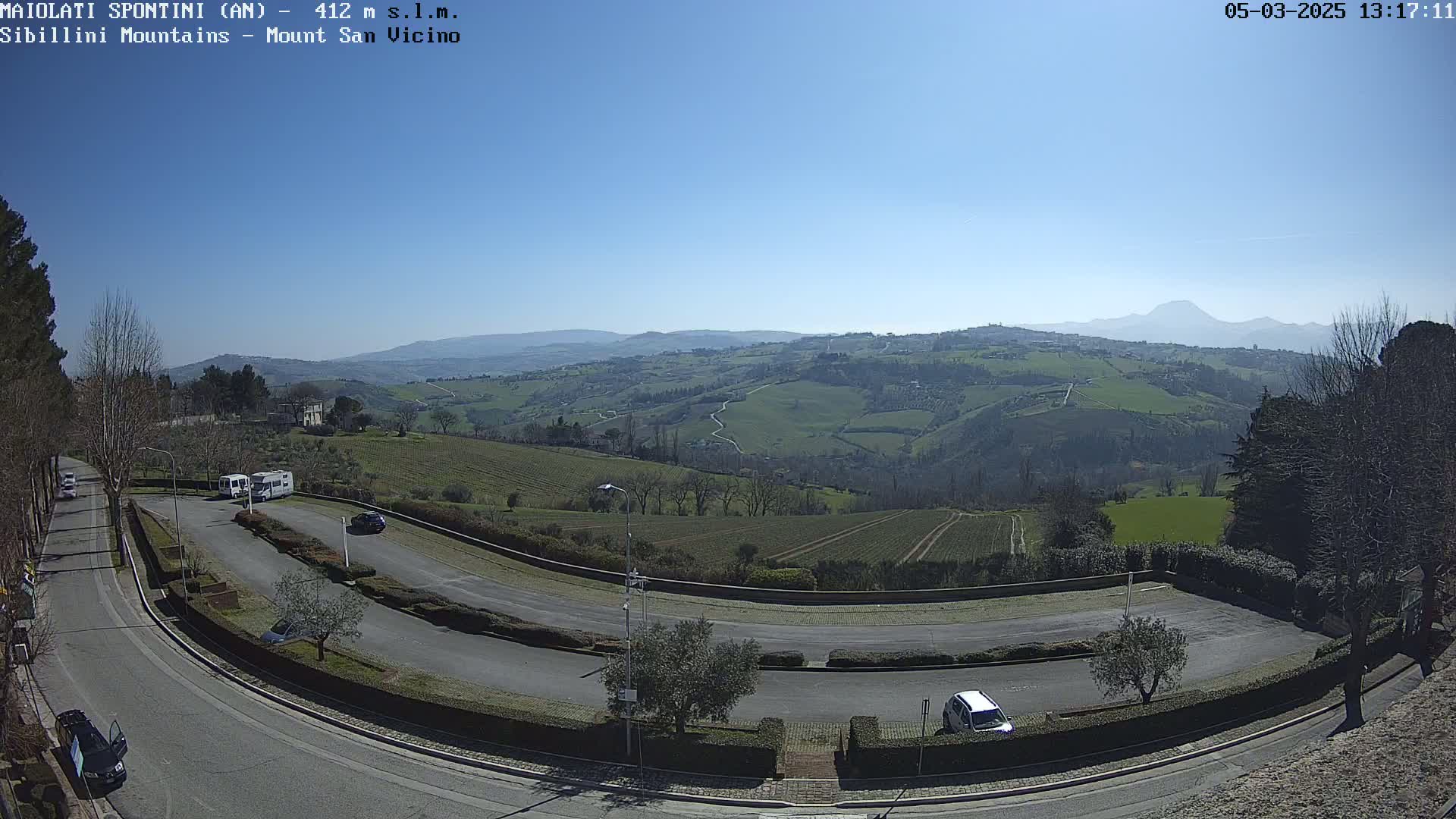 A sunny, clear day reveals a sweeping vista of rolling green hills and a parking area with several vehicles, overlooked by a road curving through the landscape.