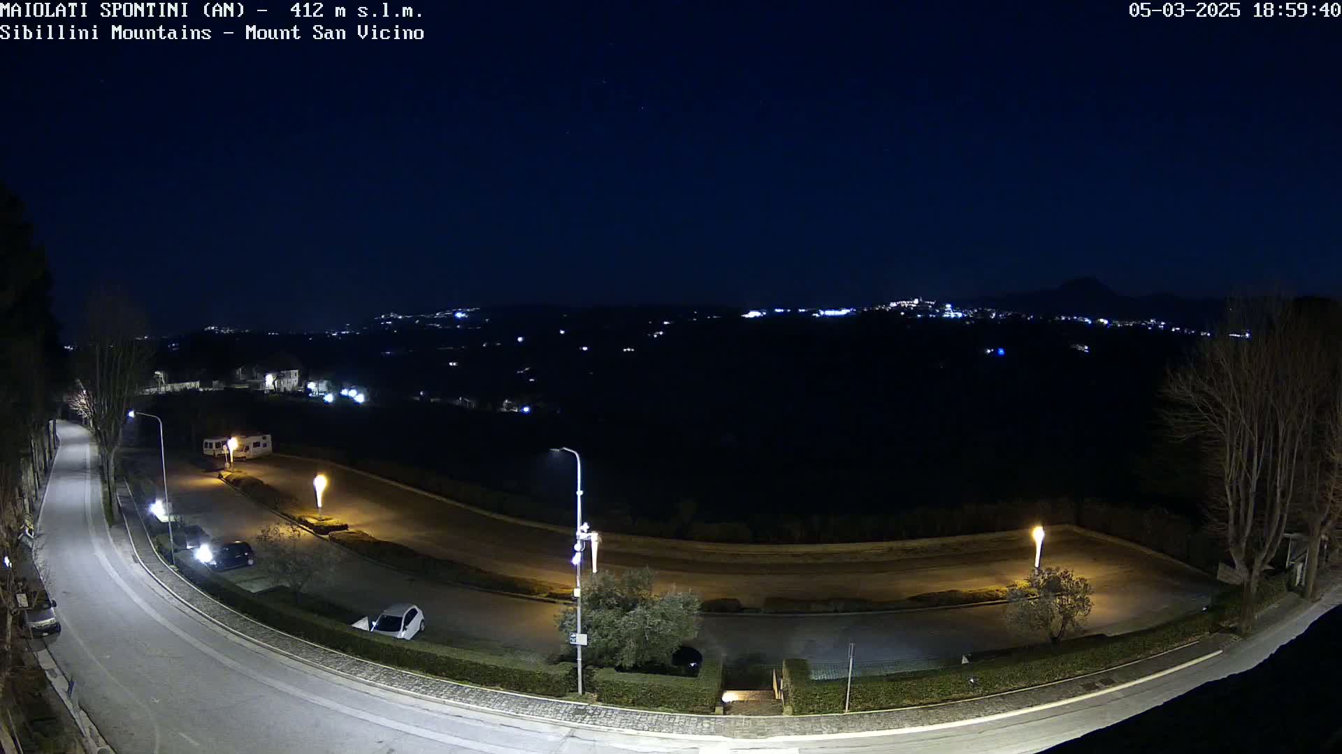 A nighttime, high-angle view of a curving road illuminated by streetlights, with a few parked cars and a distant, dimly lit town under a clear, dark sky.