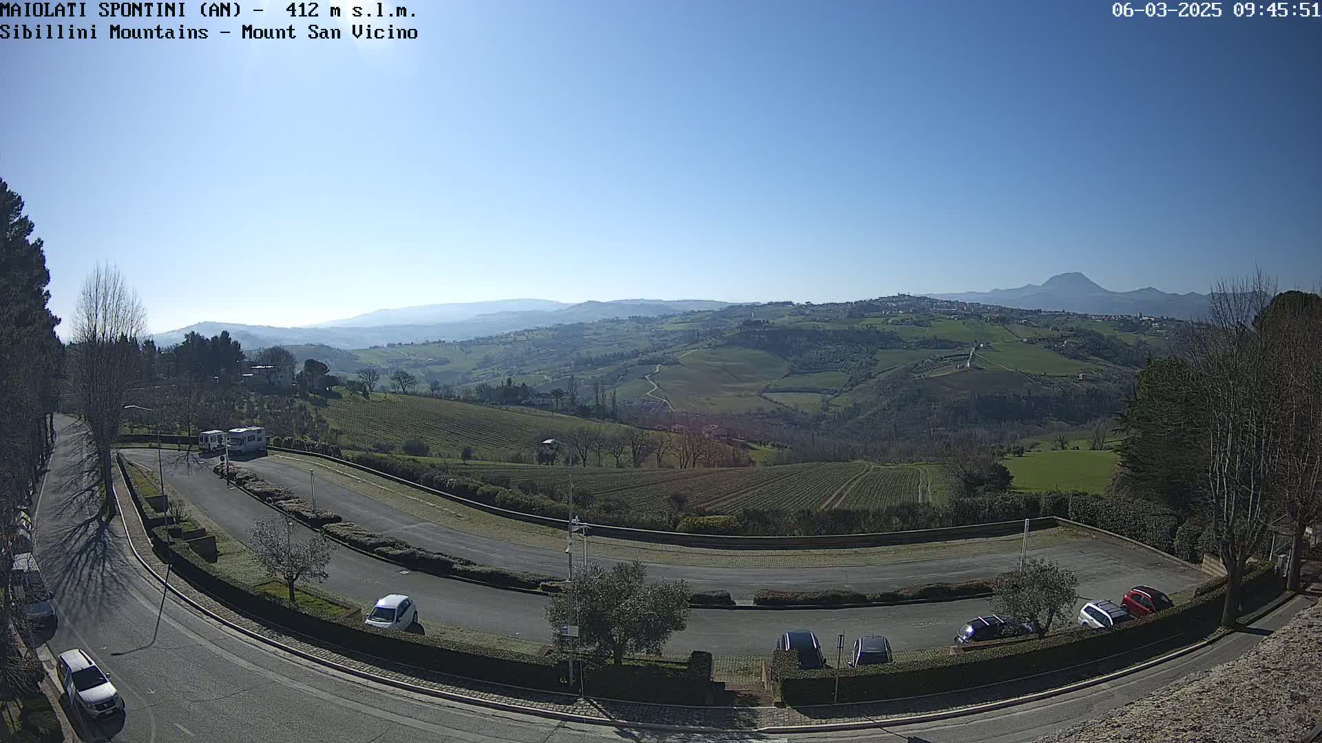 Under a clear blue sky, a curved roadway with parked cars overlooks a hilly landscape of vineyards and distant mountains.