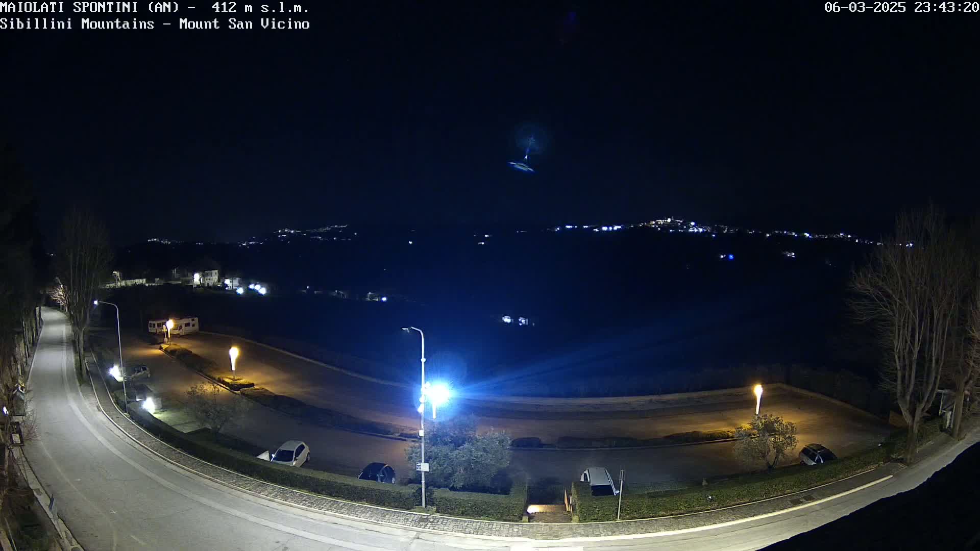 A nighttime, overhead view of a curving road with parked cars and streetlights, overlooking a distant town under a clear, dark sky.