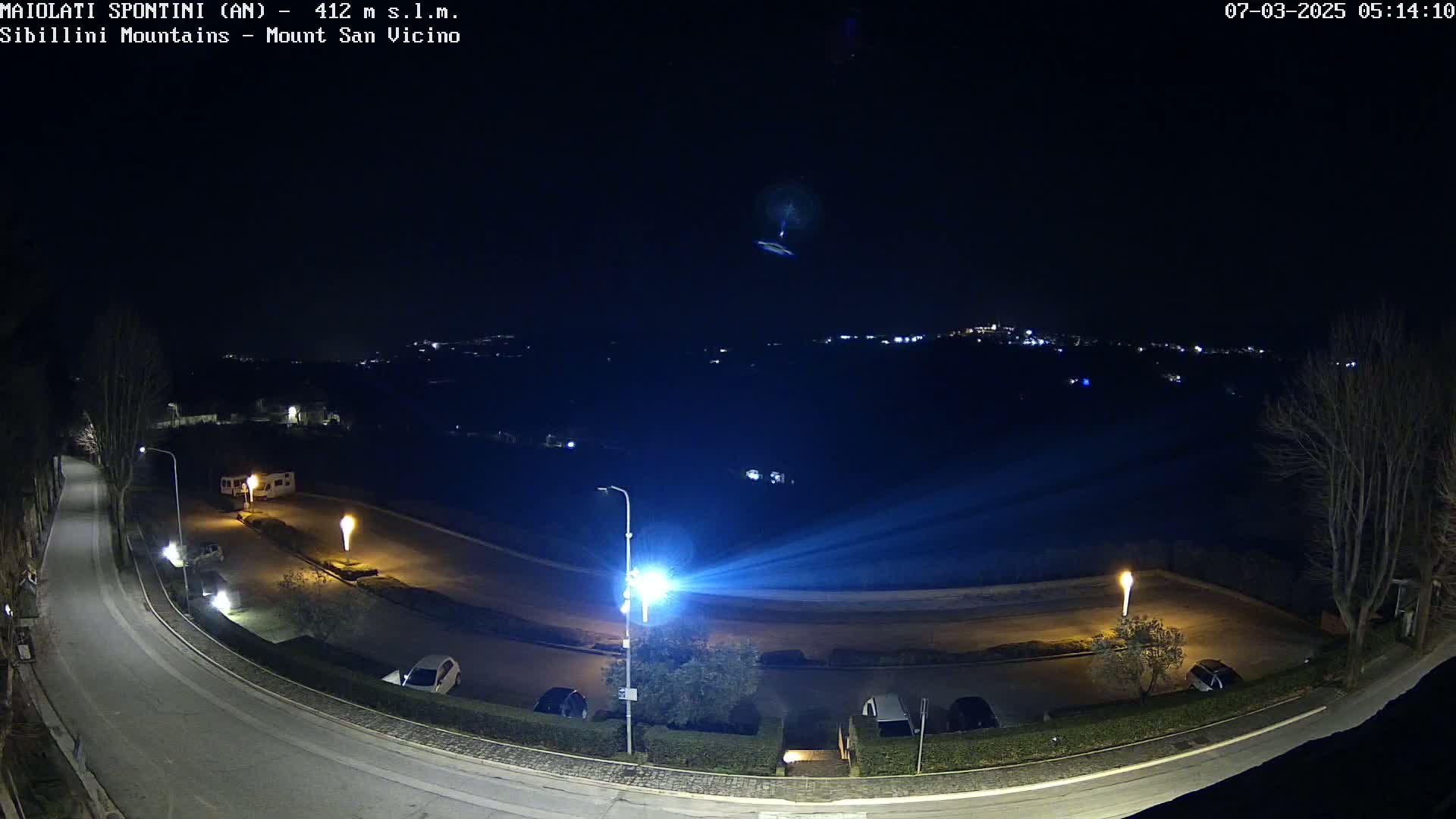 A nighttime, overhead view of a curving road with parked cars and illuminated lampposts, overlooking a distant town nestled among dark hills under a clear sky.