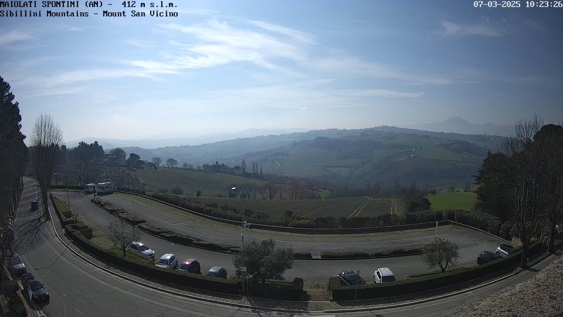 A sunny, clear day reveals a parking lot with cars, a winding road, and a view of rolling green hills and distant mountains.