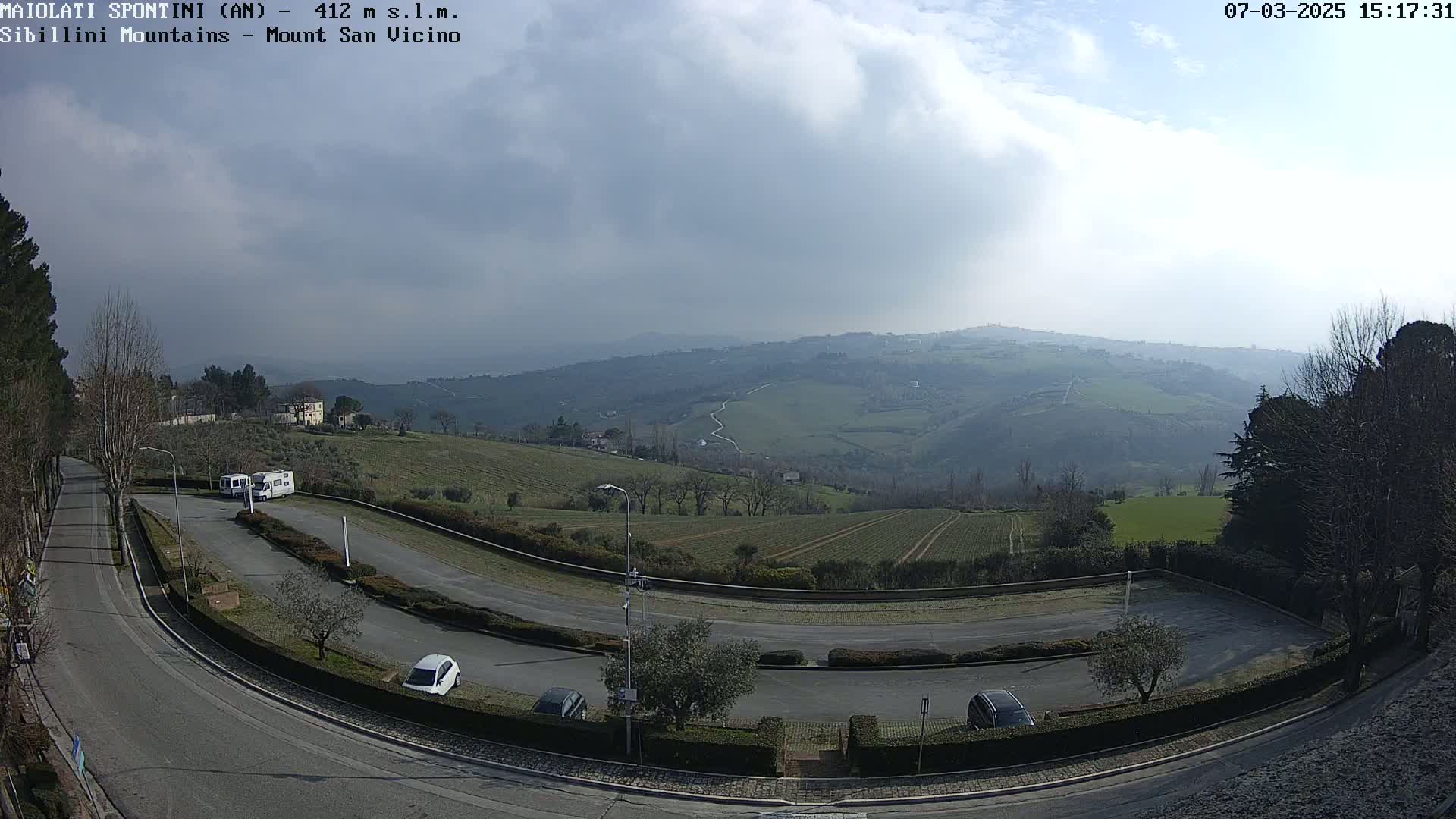 A curved road winds through a parking lot with several cars and RVs, overlooking rolling hills and farmland under a partly cloudy sky.