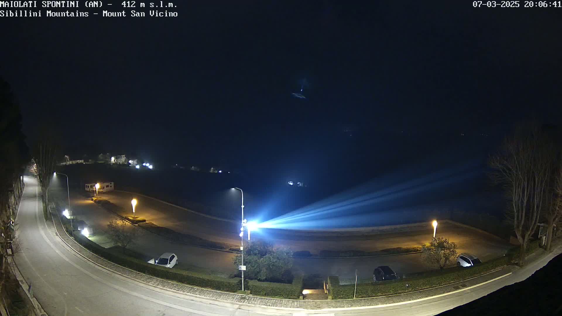 A nighttime aerial view of a curving road with parked cars and a bright light emanating from a lamppost, under a dark, clear sky.
