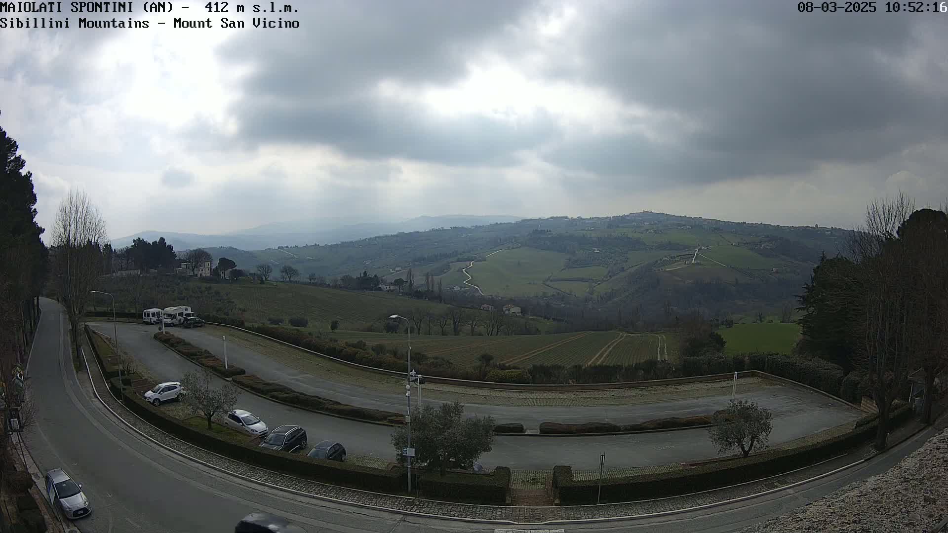 A parking lot with several cars is situated on a winding road overlooking rolling green hills under a cloudy sky.