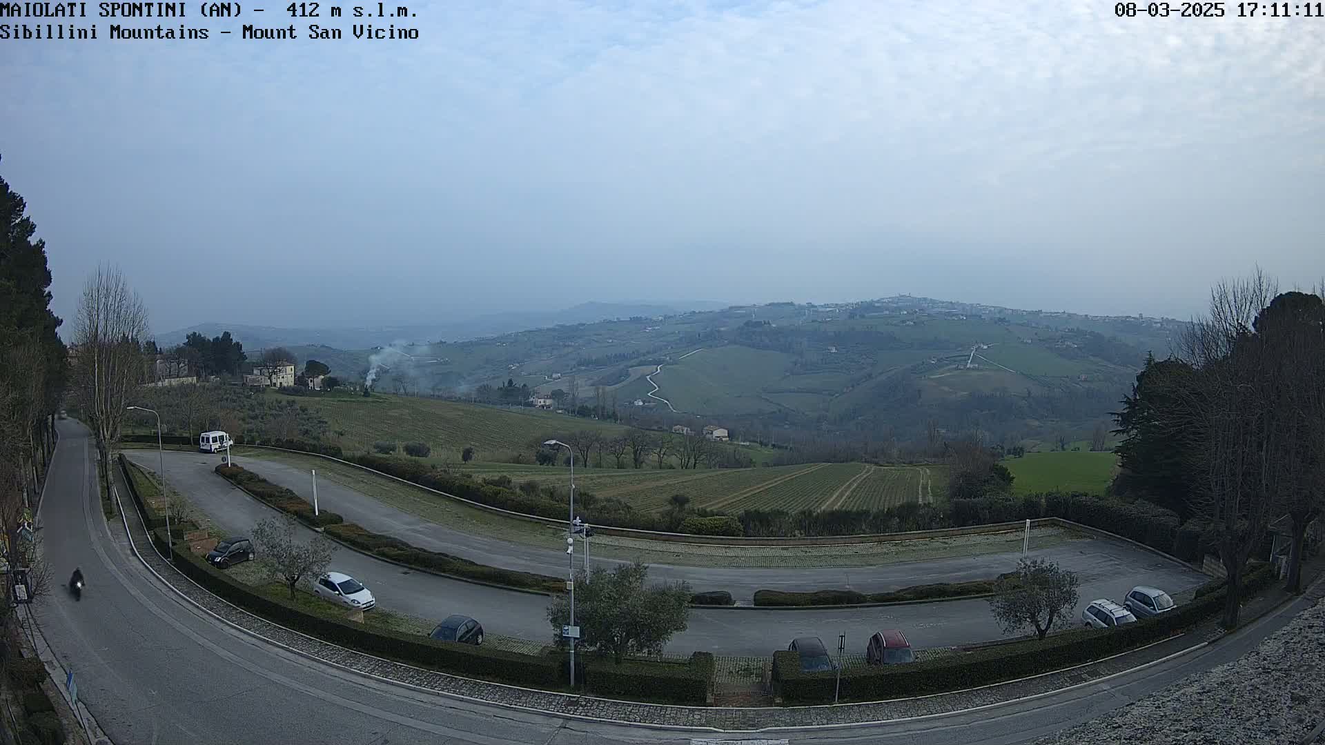 A mostly clear, overcast day reveals a winding road curving through a parking lot overlooking rolling green hills and distant buildings, with several cars parked.