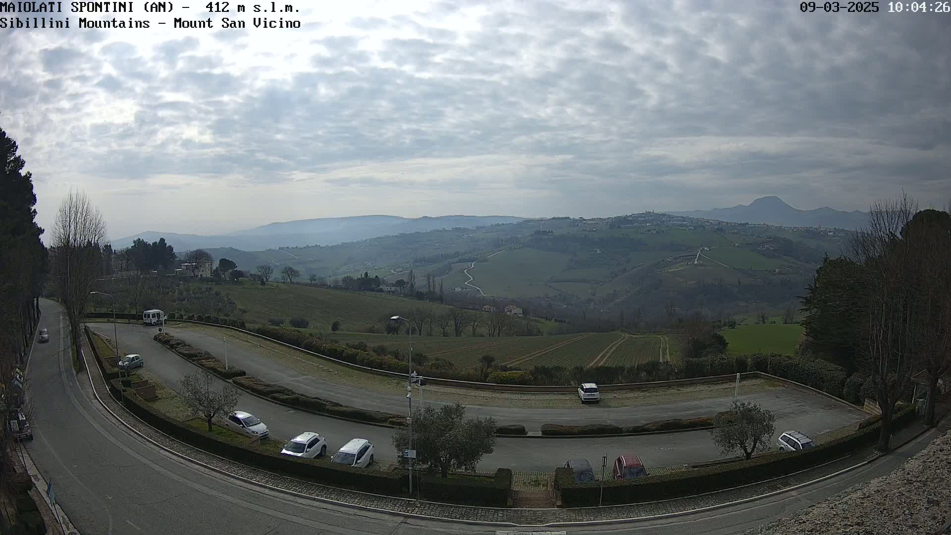 A winding road curves through a parking lot overlooking a hilly landscape under a cloudy sky, with several cars parked.