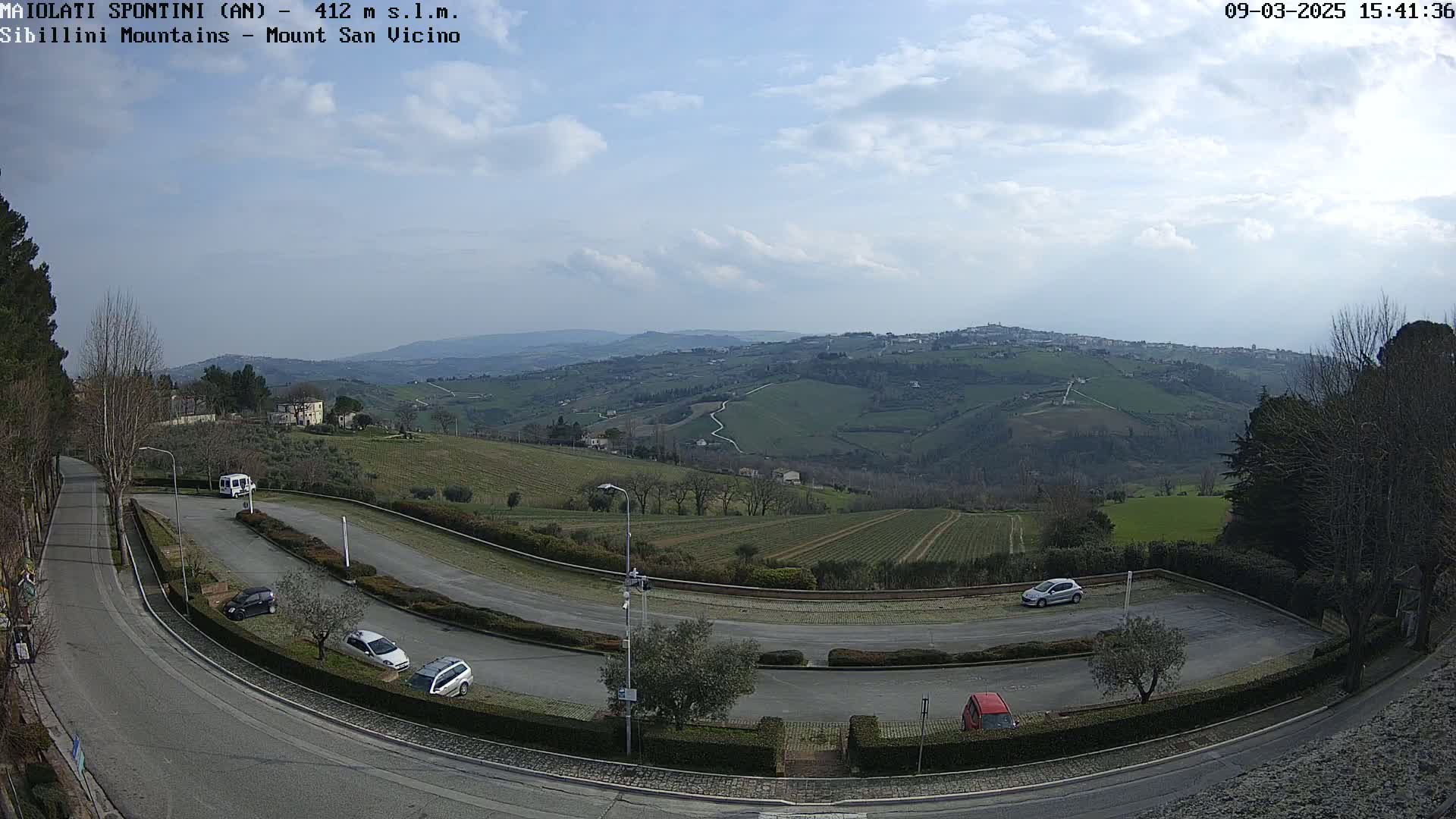 A mostly sunny, partly cloudy day overlooks a winding road and a parking lot with several cars, situated amongst rolling green hills and vineyards.