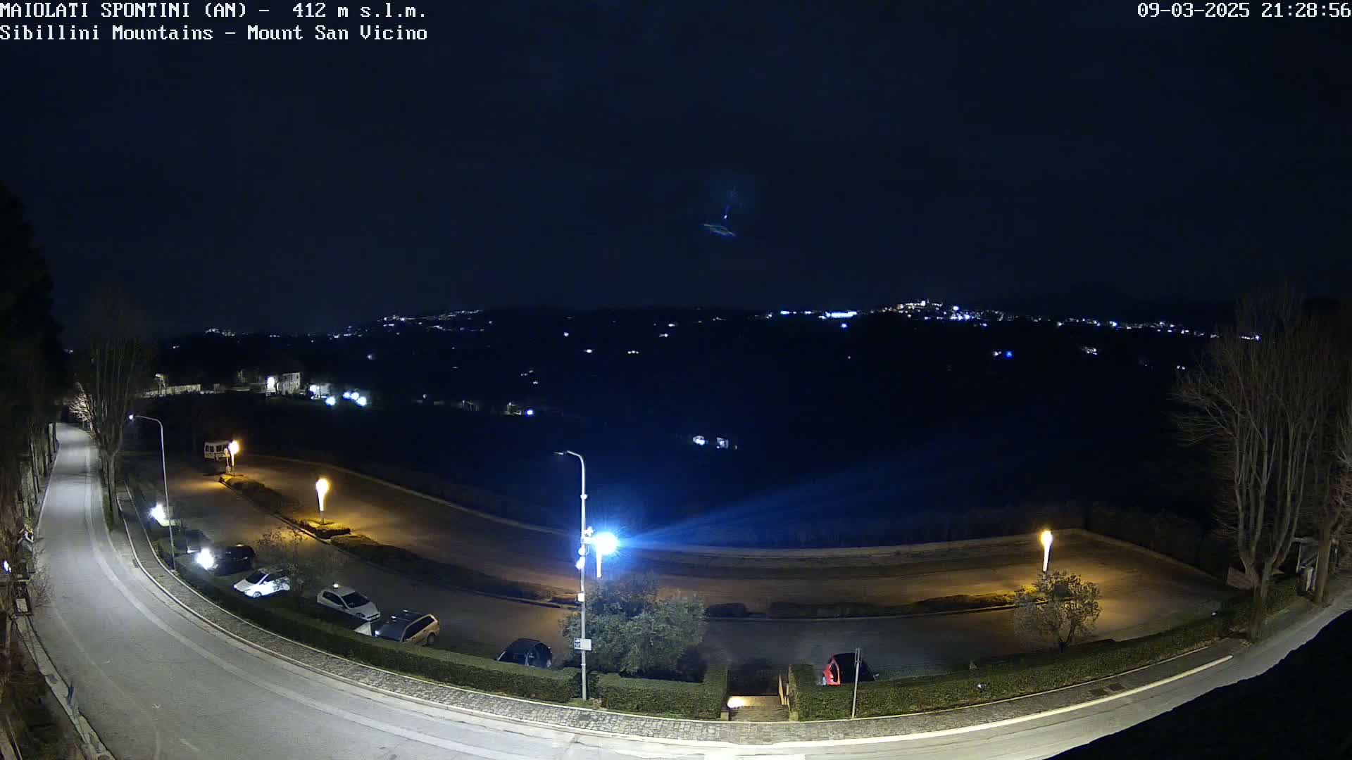 A nighttime, high-angle view of a curving road with parked cars and illuminated streetlights, overlooking a distant town under a dark sky with a faint light streak visible.