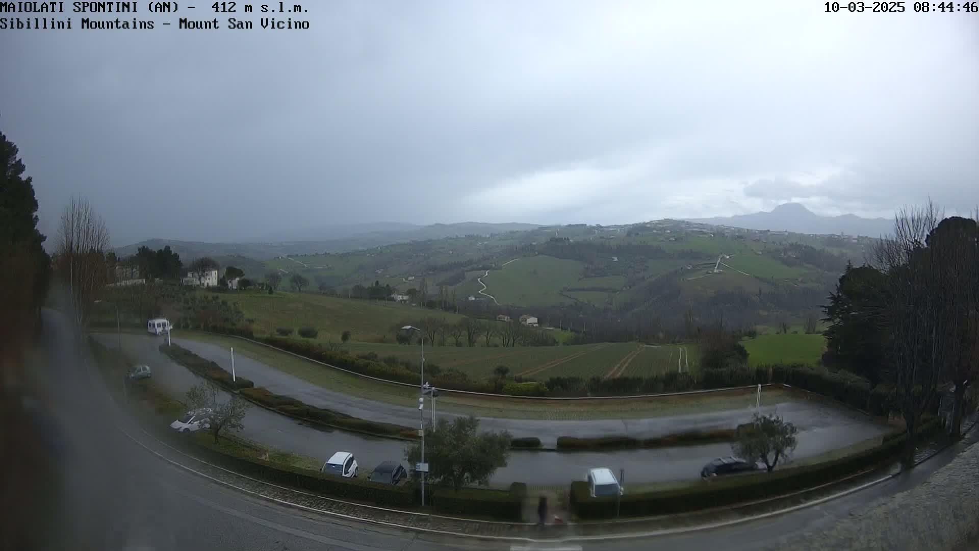 A wet, overcast view of a curving road and several parked cars overlooks a hilly, green landscape under a gray sky.