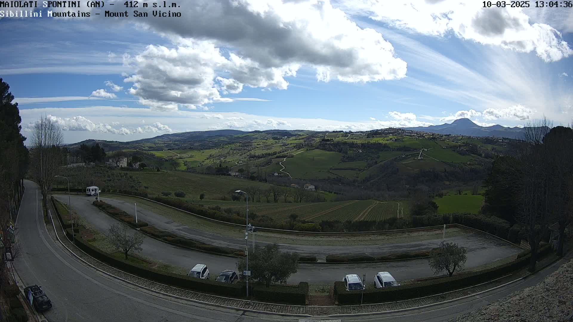 A partly cloudy day reveals a hilly landscape with vineyards and a parking lot containing several cars, overlooking a mountain range in the distance.