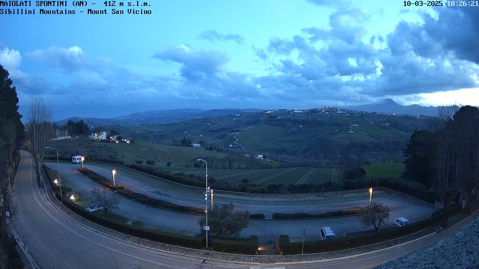 A high-angle, twilight view of a curved roadway in a parking lot overlooking rolling hills and distant mountains under a partly cloudy sky.