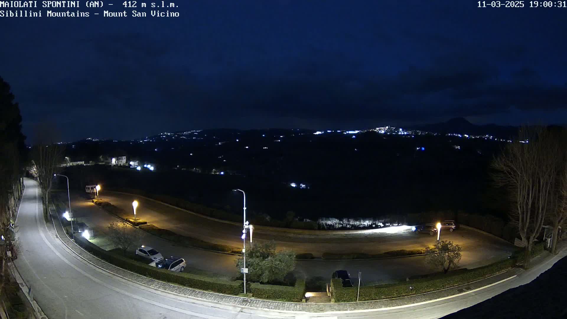 A nighttime view of a winding road with parked cars, illuminated by streetlights, overlooks a dark, distant city under a clear, dark sky.