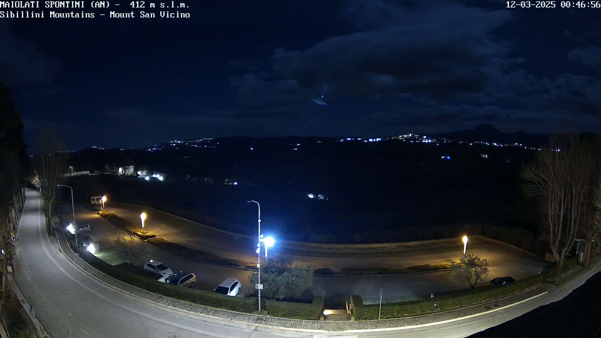 A nighttime, overhead view of a curving road with parked cars, illuminated by streetlights, overlooks a distant town nestled in a dark landscape under a partly cloudy sky.
