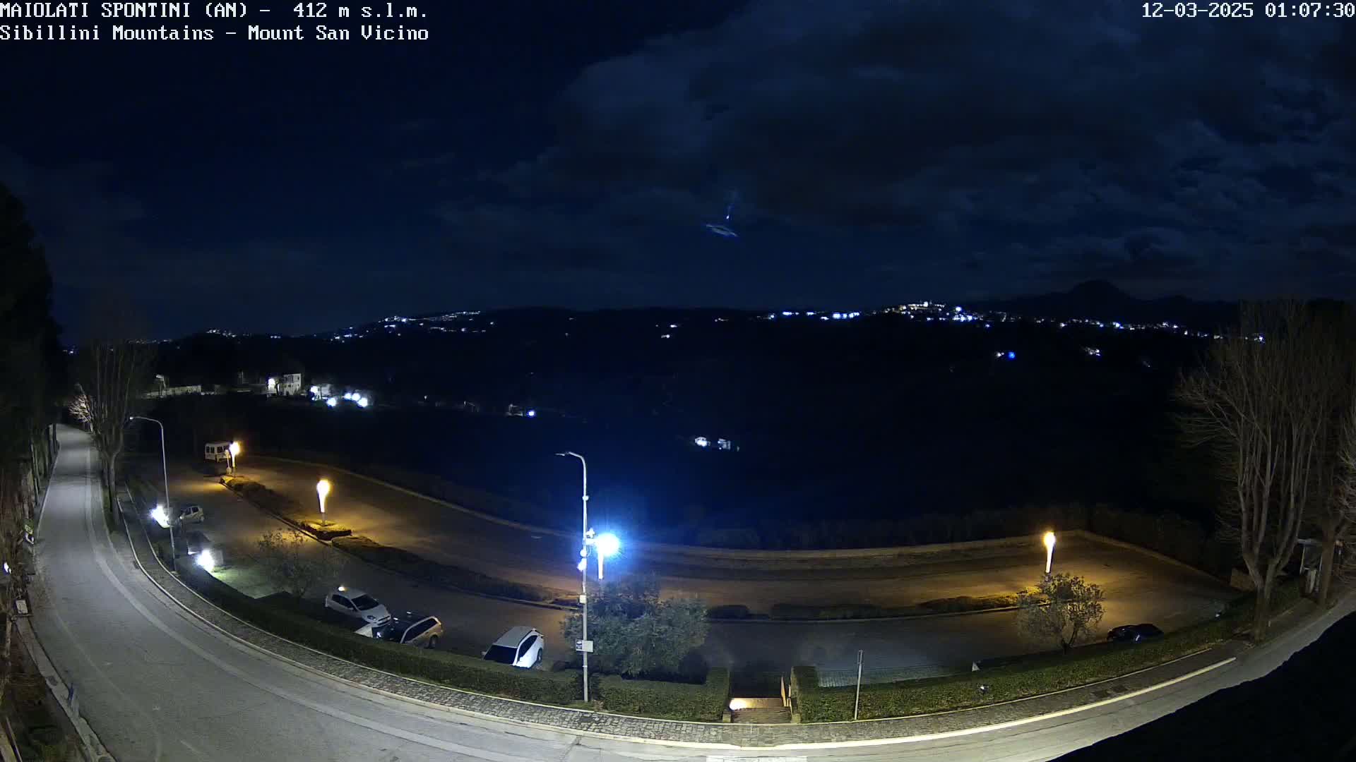 A nighttime, overhead view of a curving road with parked cars, illuminated by streetlights, overlooking a distant town under a partly cloudy sky.