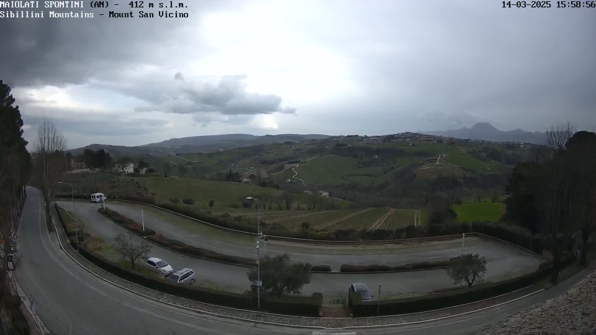 A winding road curves through a hilly landscape under an overcast sky, with a few vehicles parked along its sides and distant mountains visible in the background.