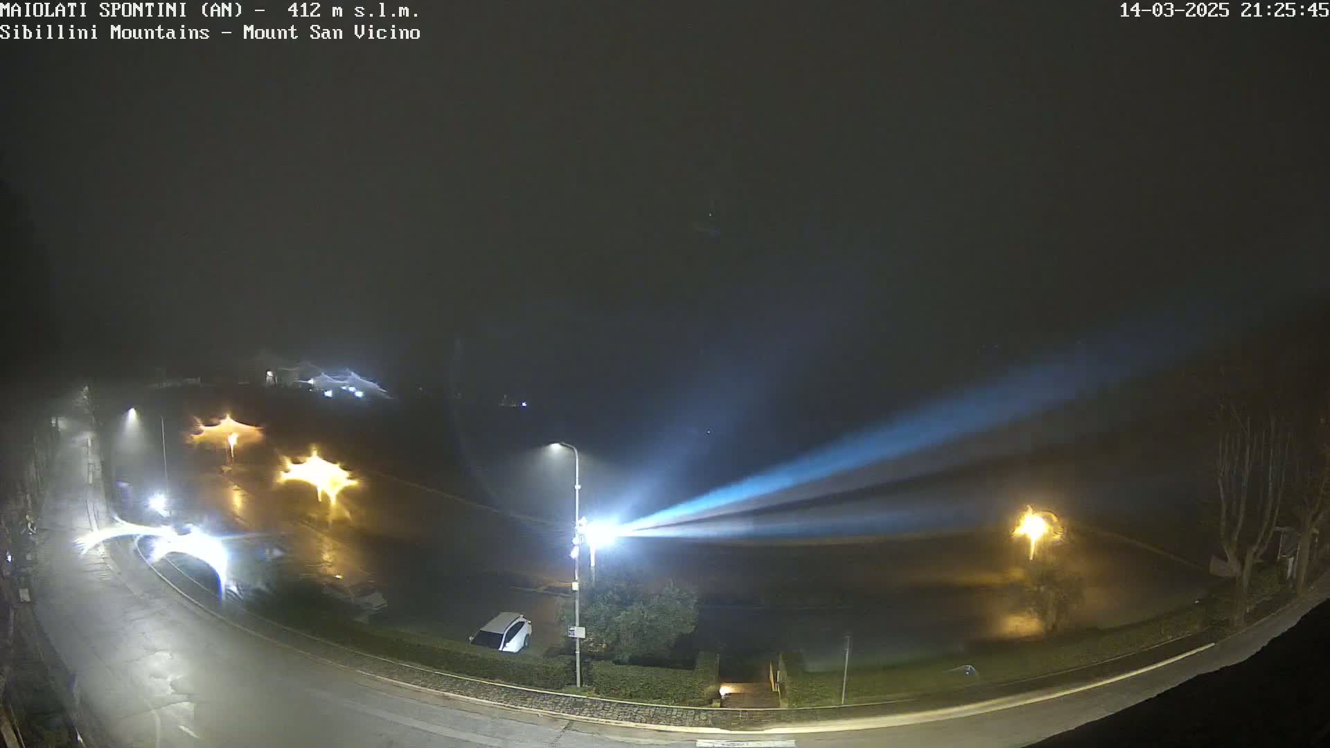 A nighttime, foggy scene shows a curved road with several illuminated streetlights and vehicles, with two bright beams of light intersecting in the distance.