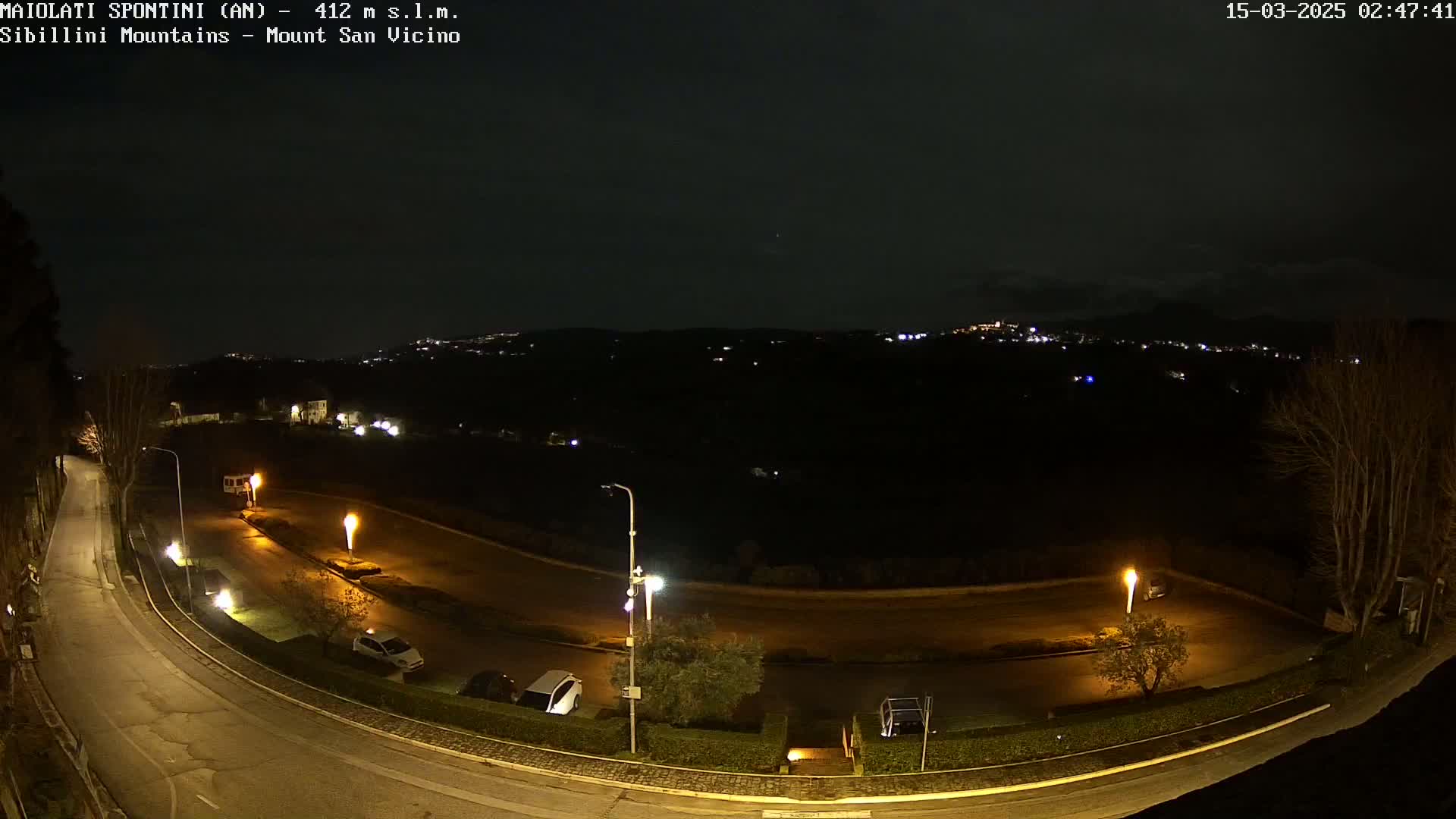 A nighttime, overhead view of a curving road illuminated by streetlights, with a few parked cars and a distant, dimly lit town under a dark sky.