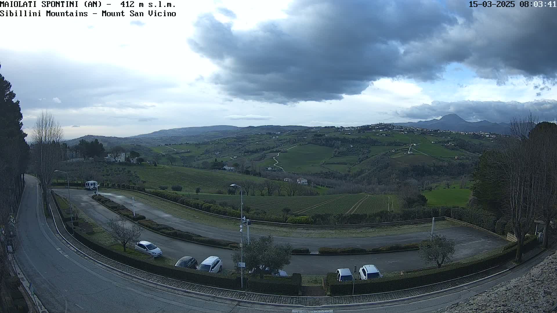 An empty parking lot curves around a hilltop overlooking a vast, rolling landscape of green fields and sparse trees under a cloudy sky.