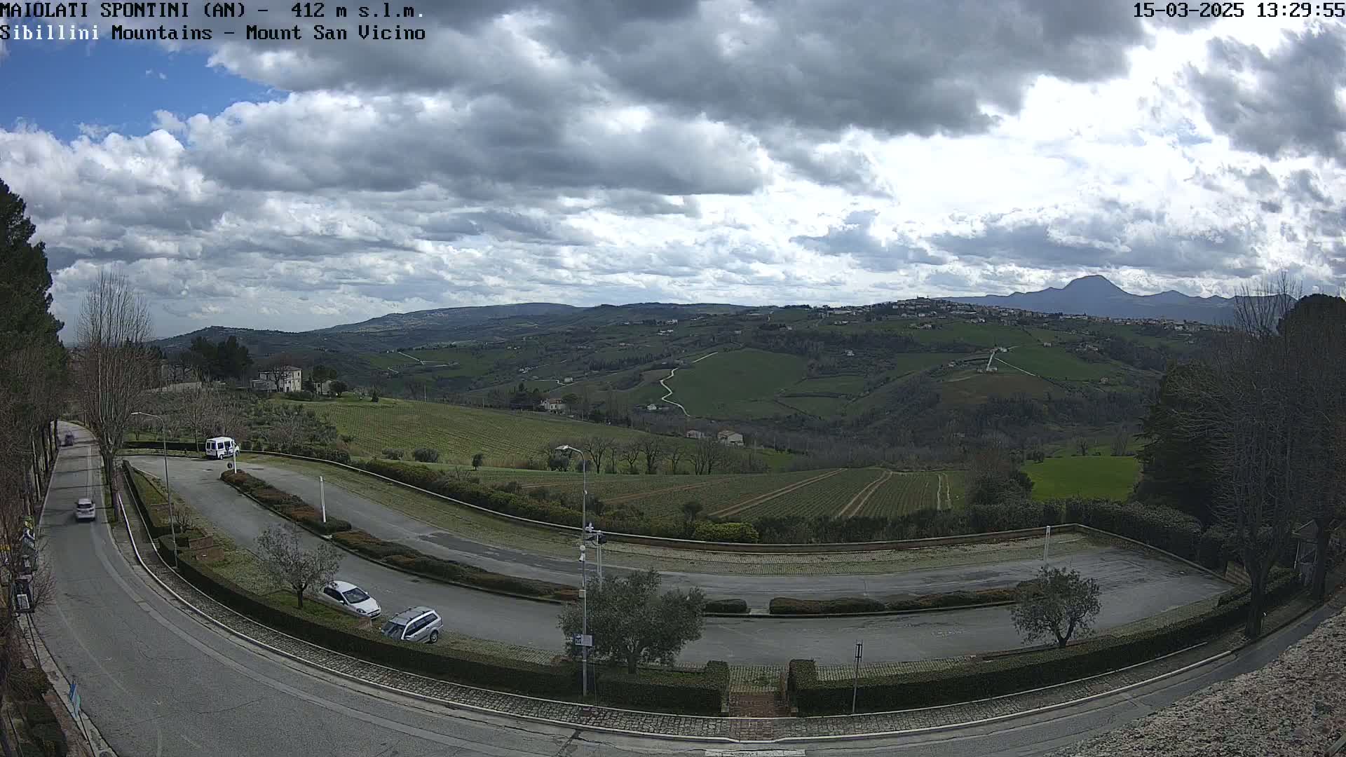 A winding road curves through a parking lot overlooking a green valley under a partly cloudy sky, with mountains in the distance.