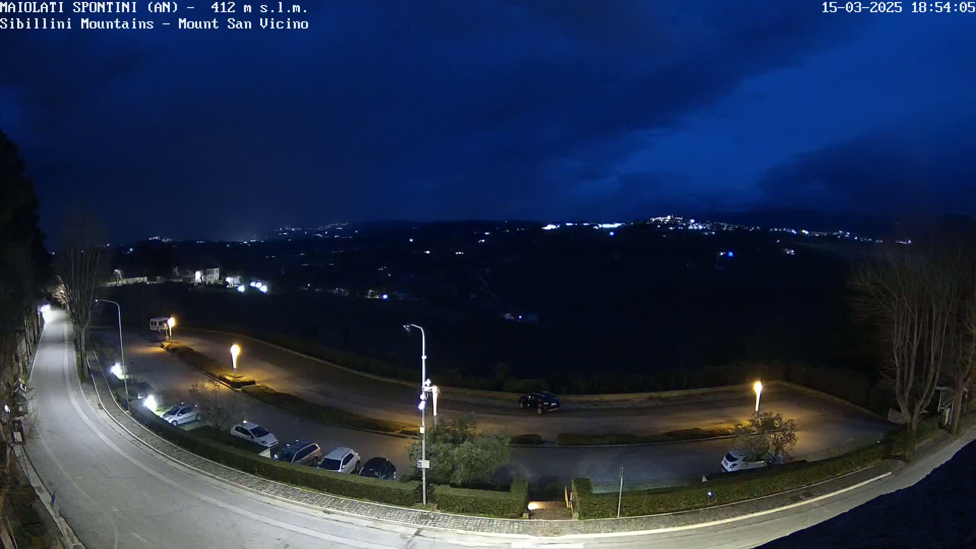 A nighttime, high-angle view shows a curving road with parked cars and streetlights, overlooking a dark, hilly landscape under a mostly clear, dark blue sky.