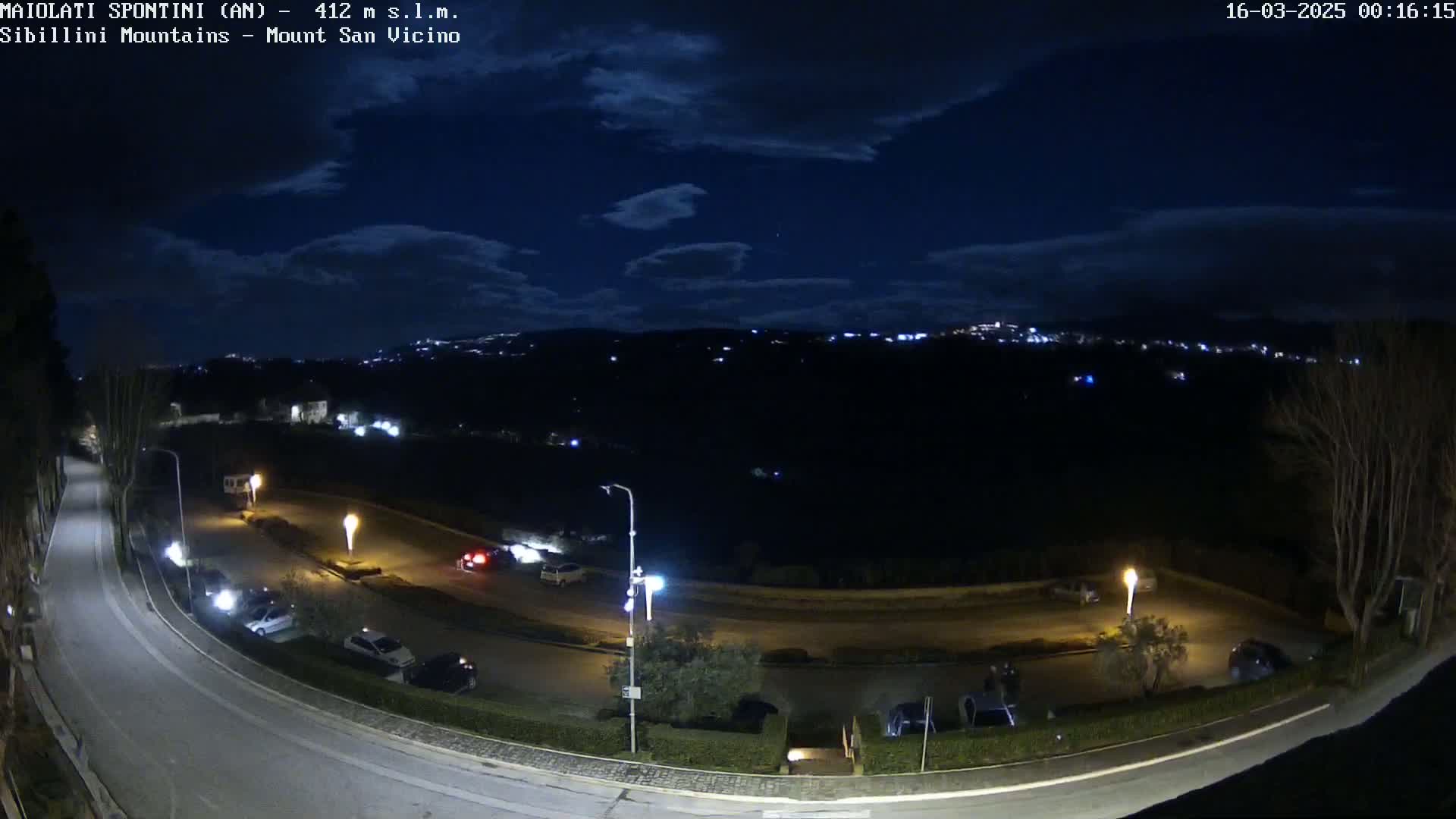 A nighttime view of a curved road with parked cars and distant city lights under a partly cloudy sky.