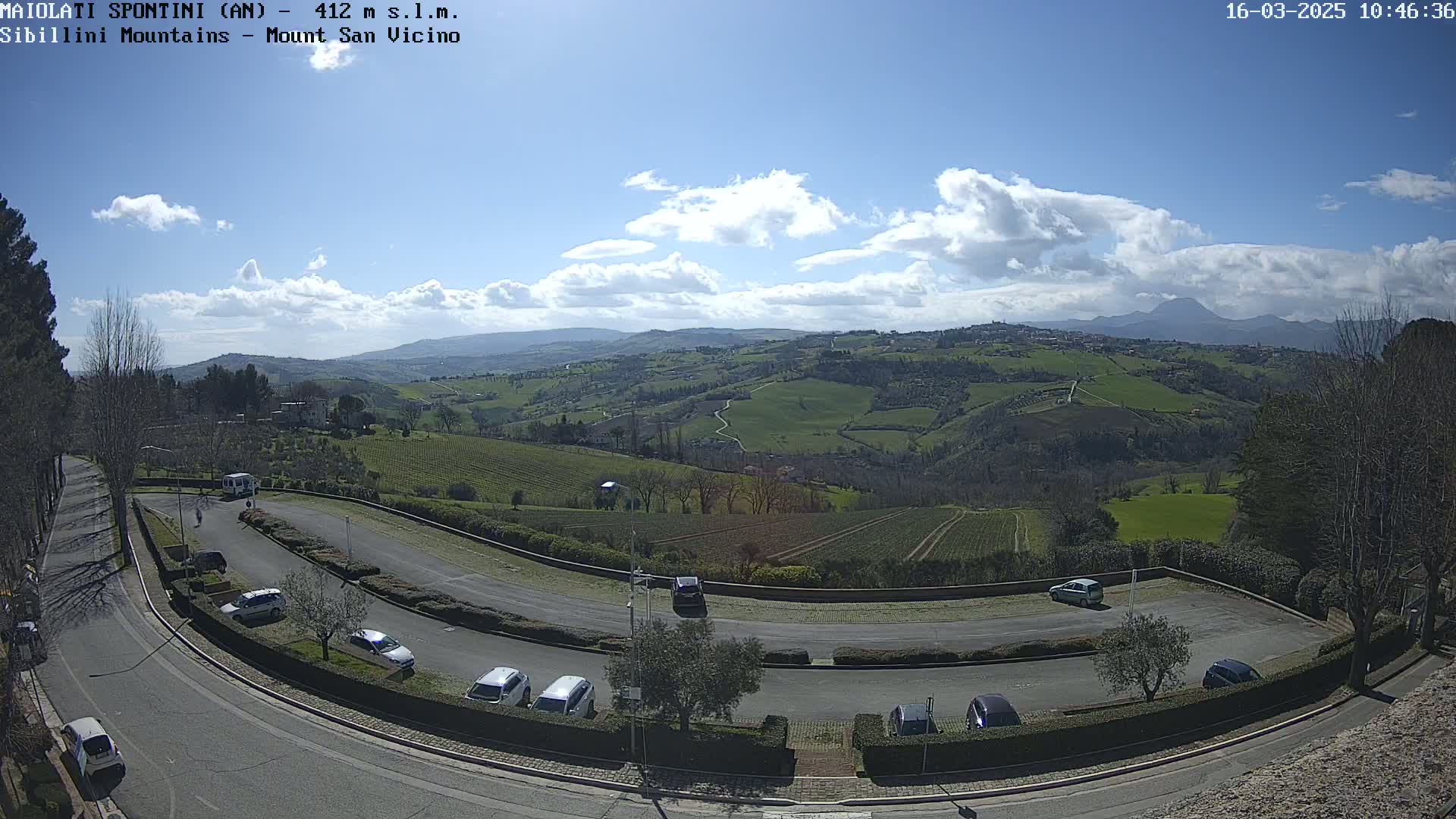 A parking lot with several cars curves around a hillside overlooking a vast, rolling green landscape under a partly cloudy blue sky.