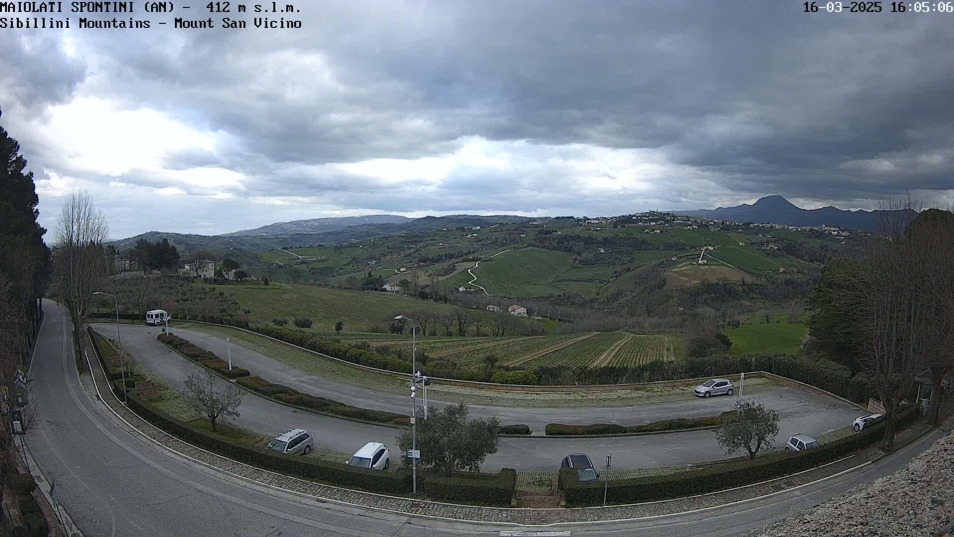 A winding road curves through a parking lot overlooking rolling green hills and vineyards under a cloudy sky.
