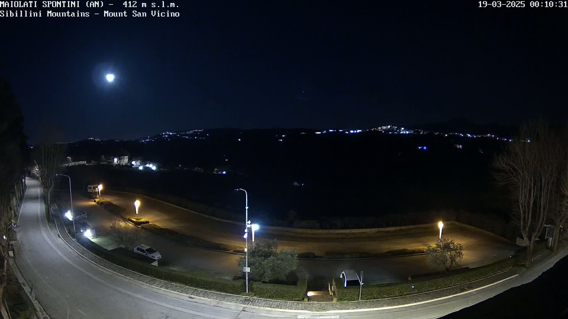 A nighttime view of a curving road illuminated by streetlights, with a bright moon in the sky and distant city lights visible on a hillside.
