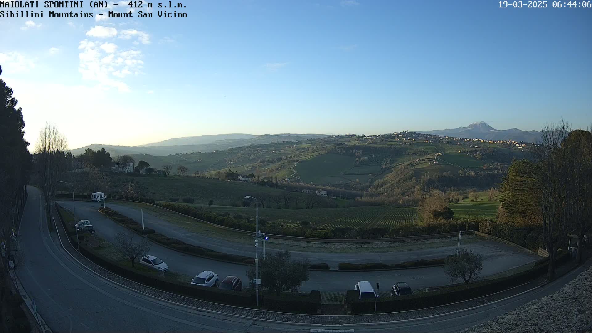 A clear, sunny day overlooks a hilly landscape with a parking lot and winding road in the foreground, and distant mountains in the background.