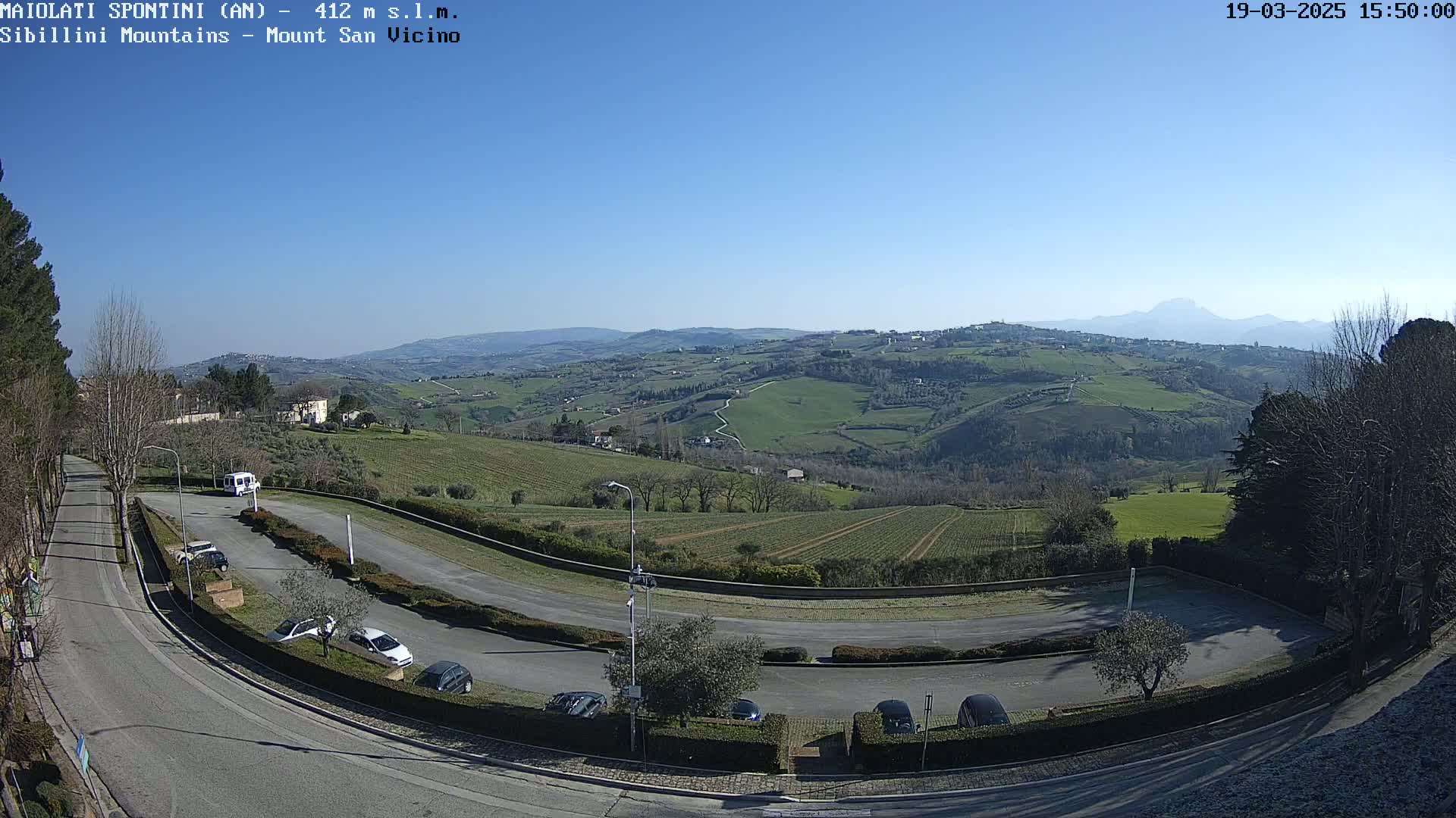 A sunny, high-angle view shows a paved parking lot and curving road nestled among green hills and farmland, with mountains visible in the distance.