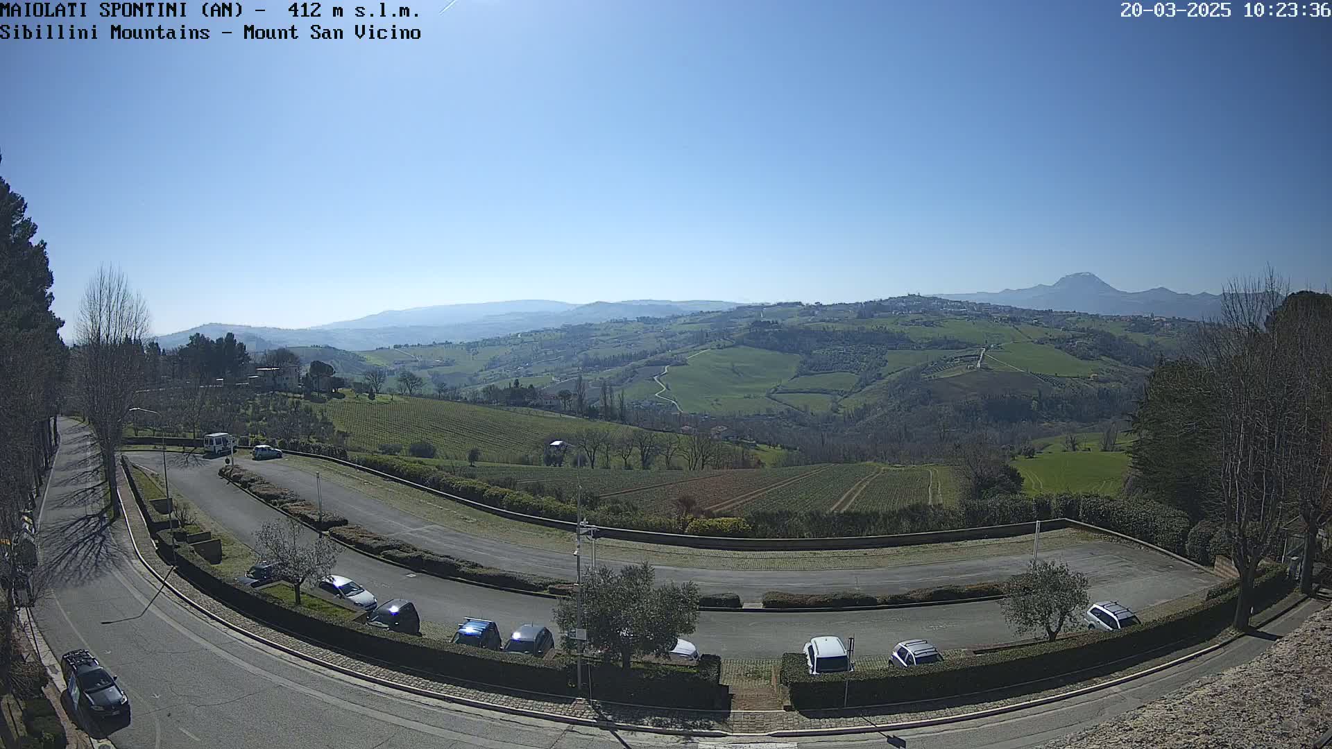 A sunny, outdoor view of a curved parking lot and roadway overlooking a vast, rolling green landscape with distant mountains.