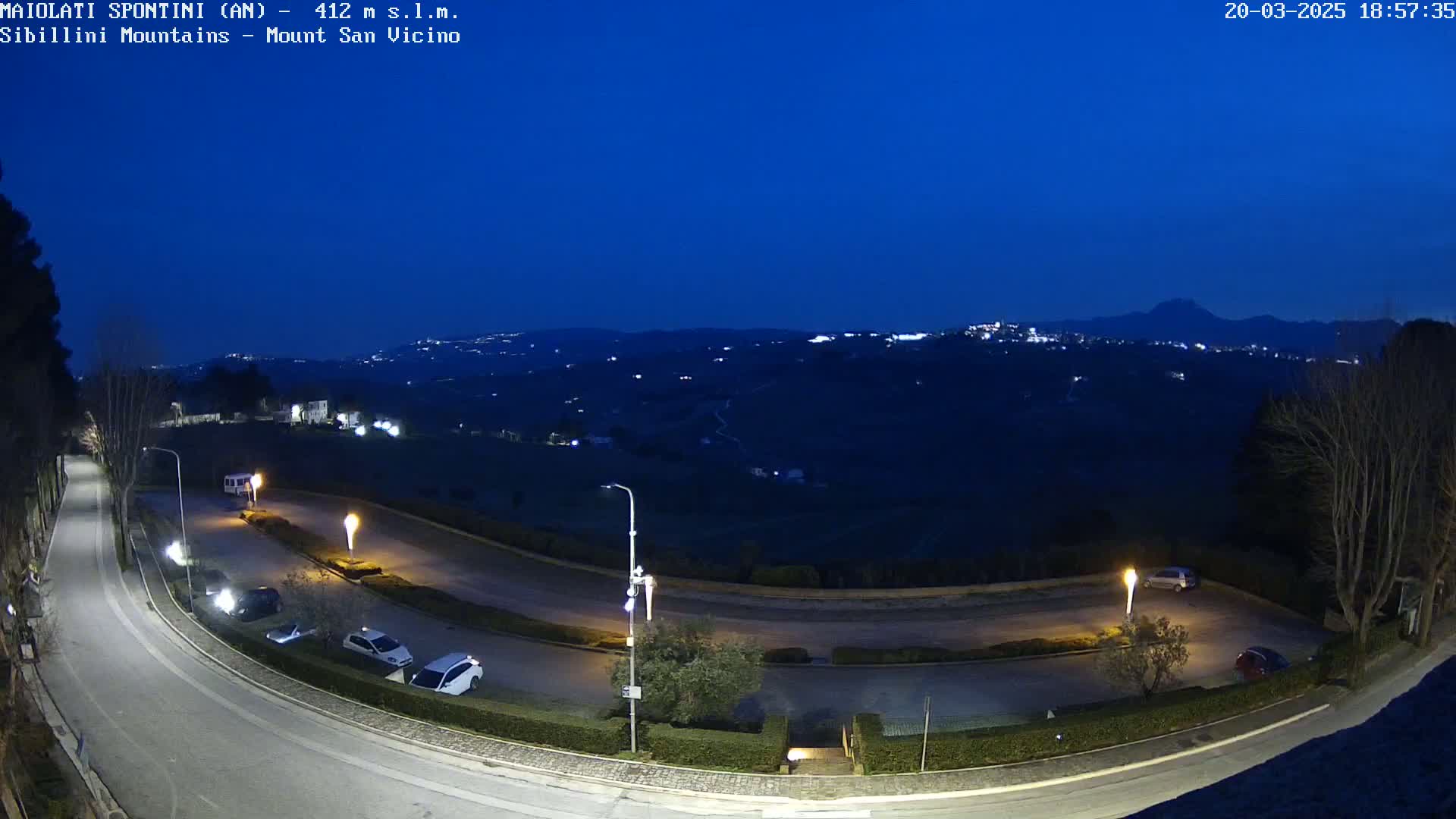 A nighttime, overhead view of a curving road with parked cars, streetlights, and a distant, dark mountain range under a clear, deep blue twilight sky.