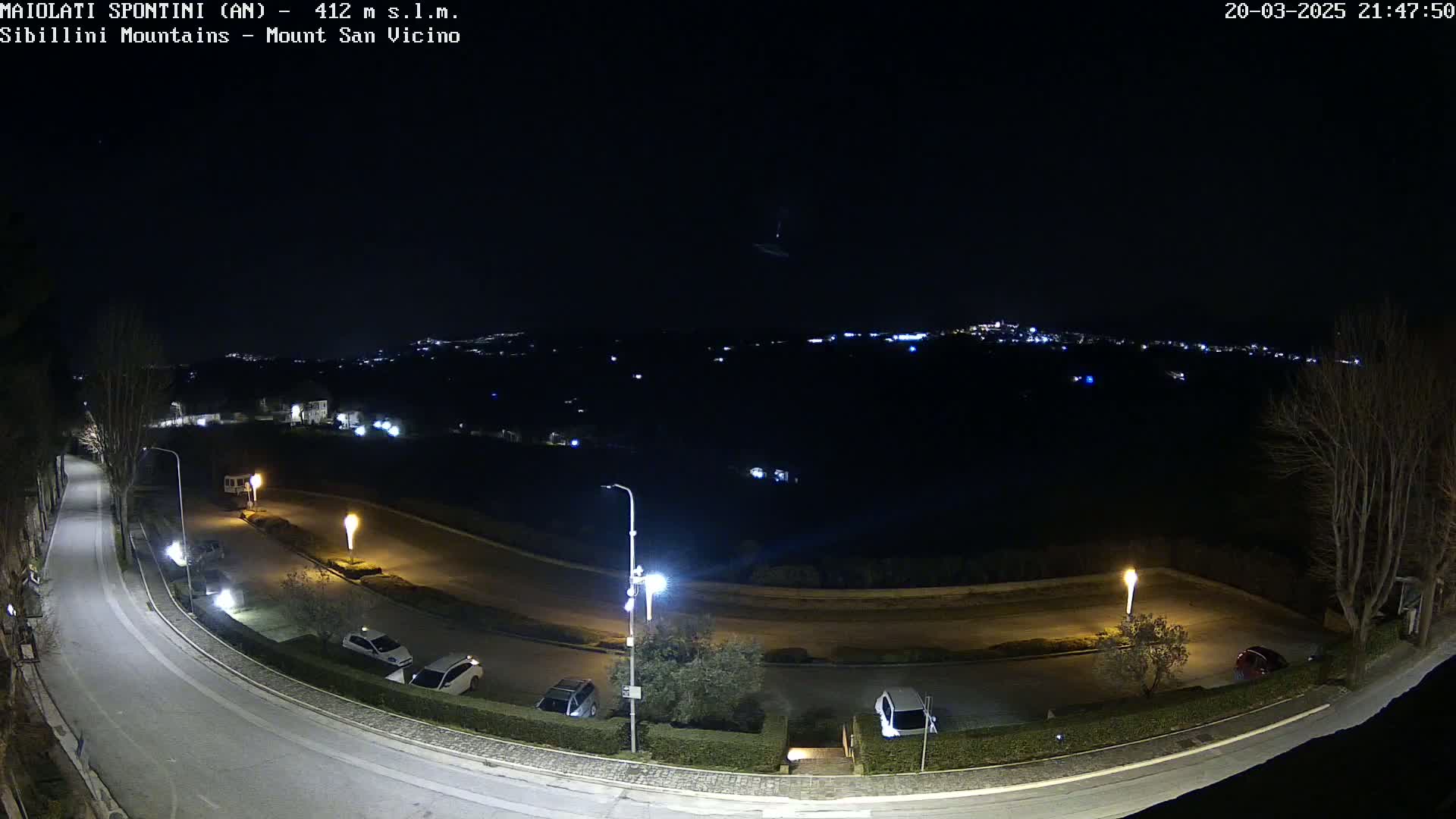 A nighttime, high-angle view of a curving road with parked cars and illuminated streetlights, overlooking a distant town nestled among dark hills under a clear sky.