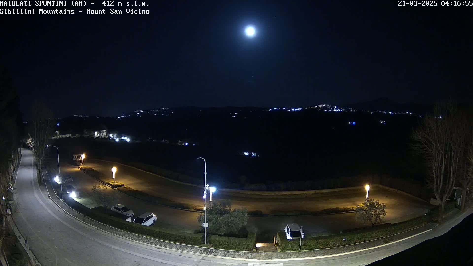 A bright moon illuminates a nighttime scene of a curving road with parked cars and streetlights, overlooking a distant town nestled in dark hills.